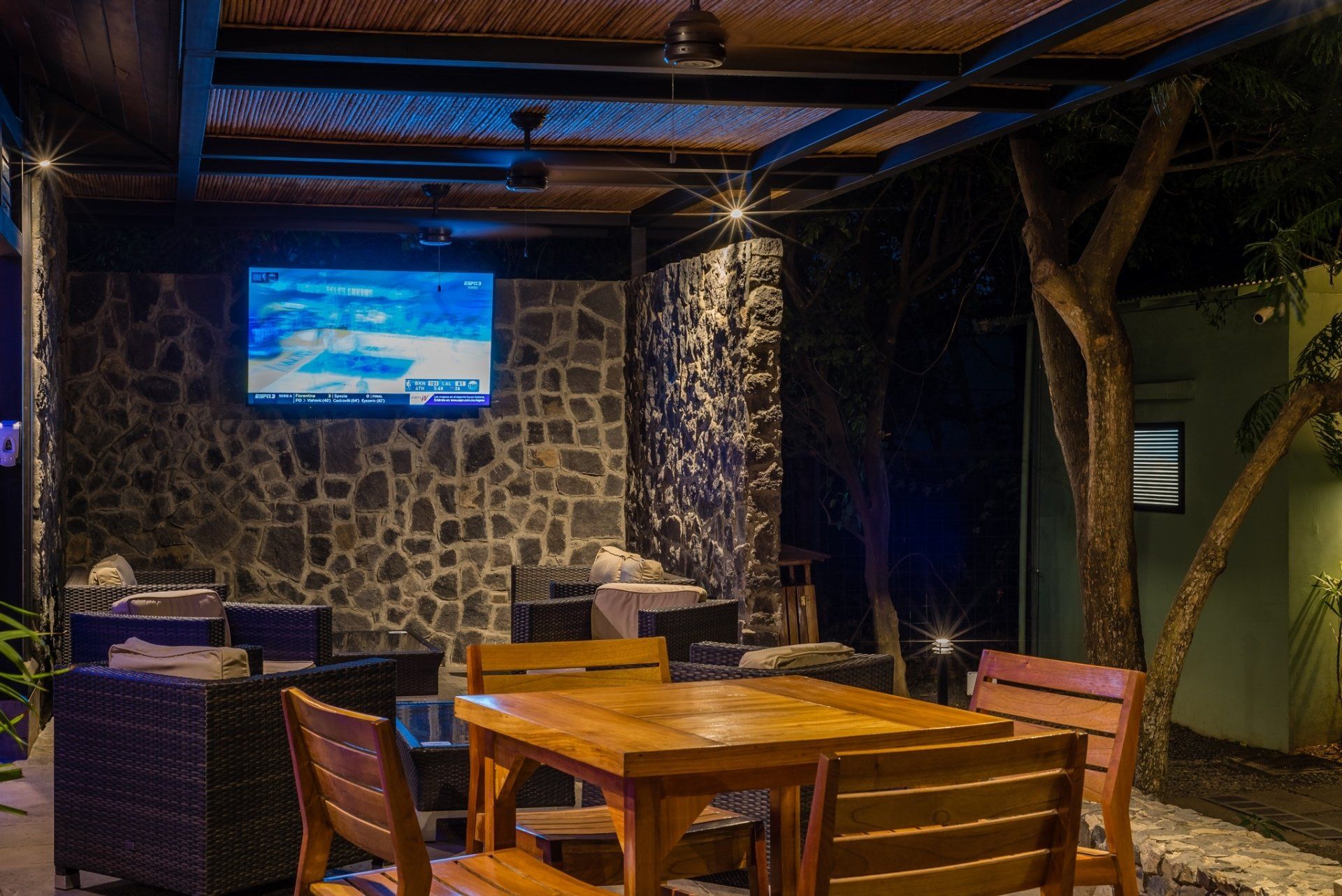 Outdoor patio dining area with wooden tables, stone walls, and a wall-mounted TV at night