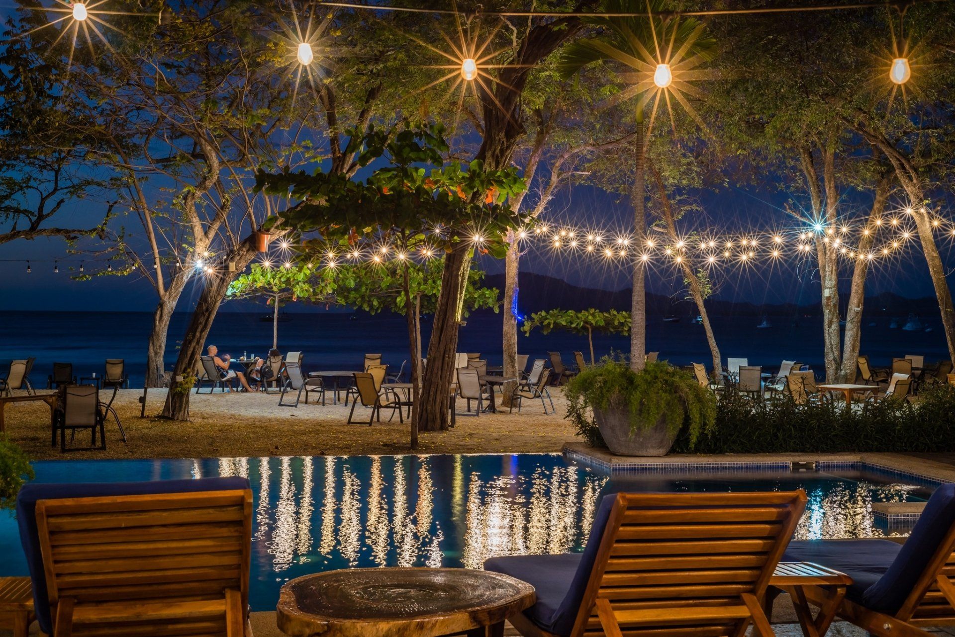 Poolside resort dining area at dusk with lit trees, ocean view, and wooden lounge chairs