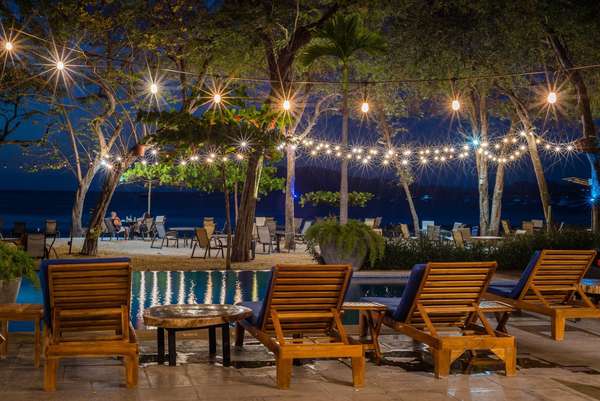 Empty beachside lounge chairs under string lights at night, with palm trees and ocean view