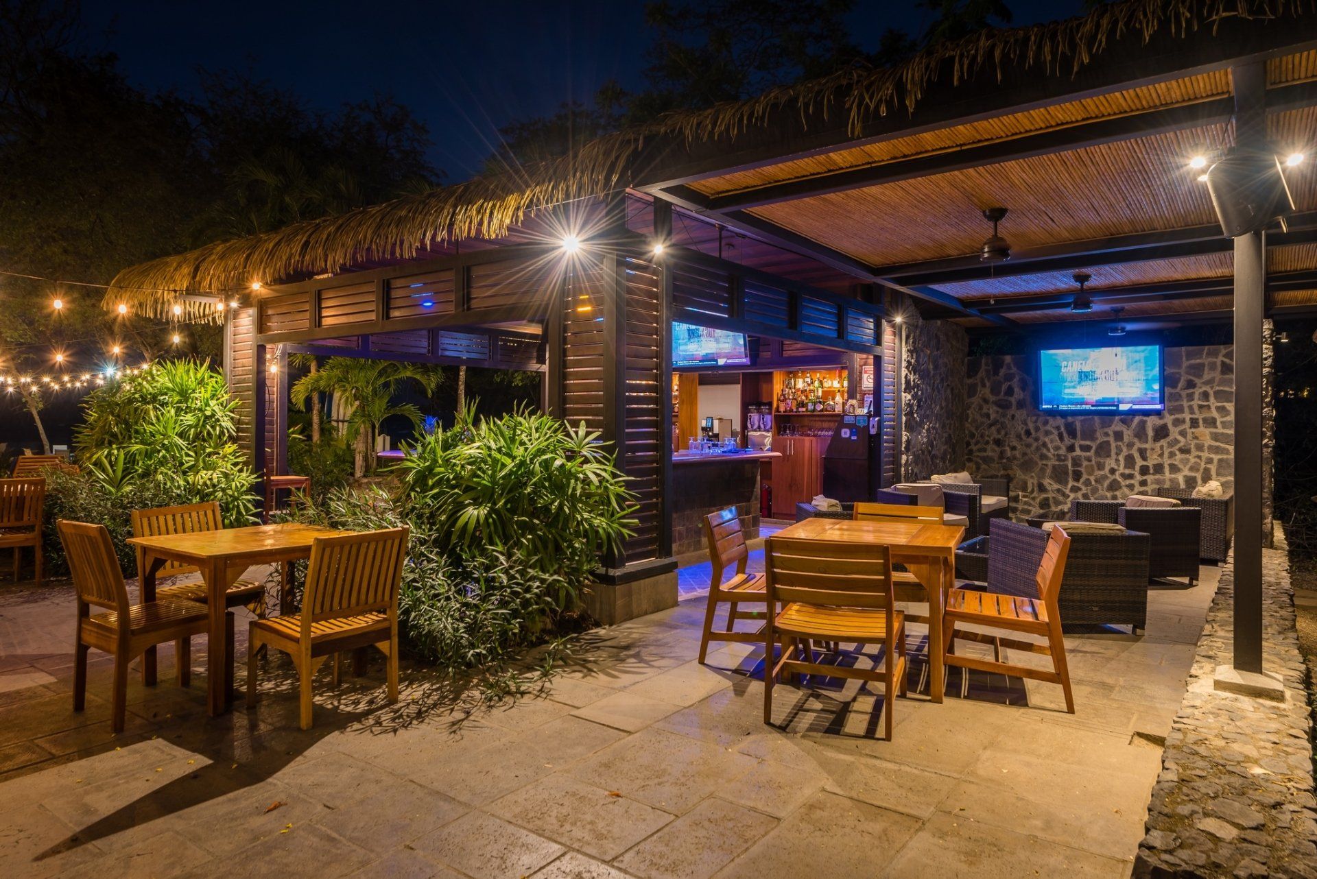 Open-air restaurant patio at night with wooden tables, warm lights, and a TV screen in the background