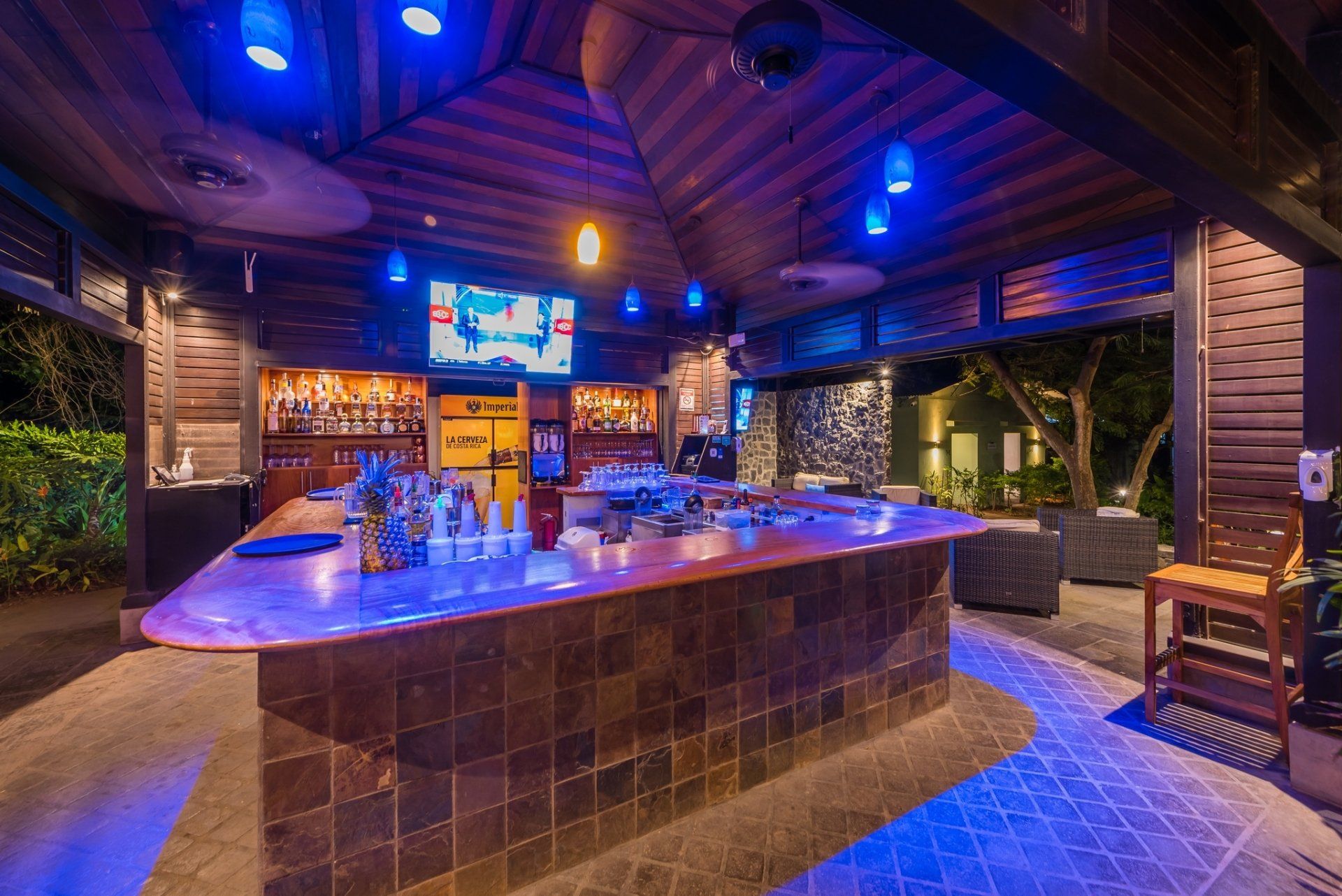 Empty modern bar interior with blue-purple lighting, tiled counter, stools, and TV screens