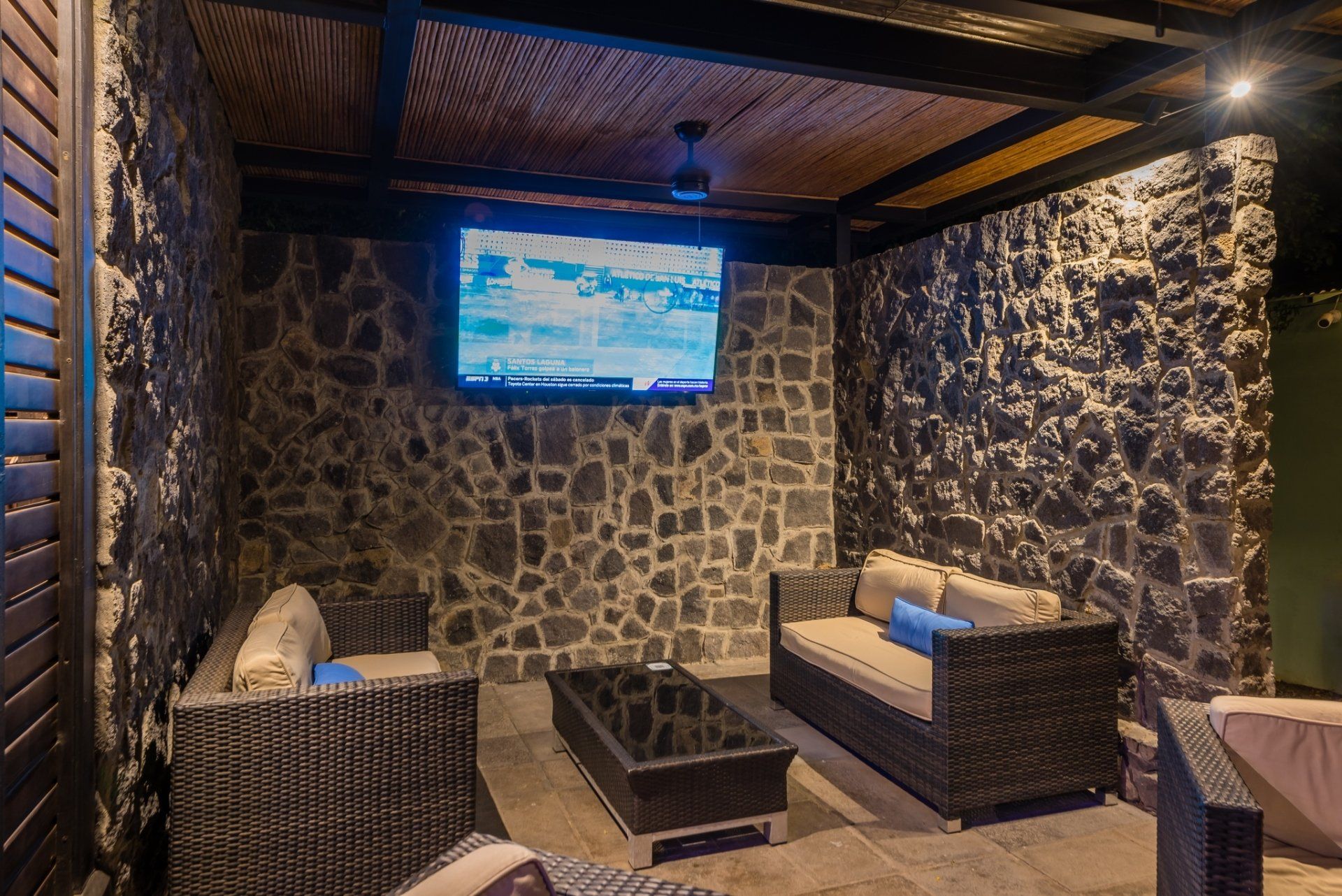 Stone-walled lounge with wicker seating, a central table, and a wall-mounted TV under warm lighting