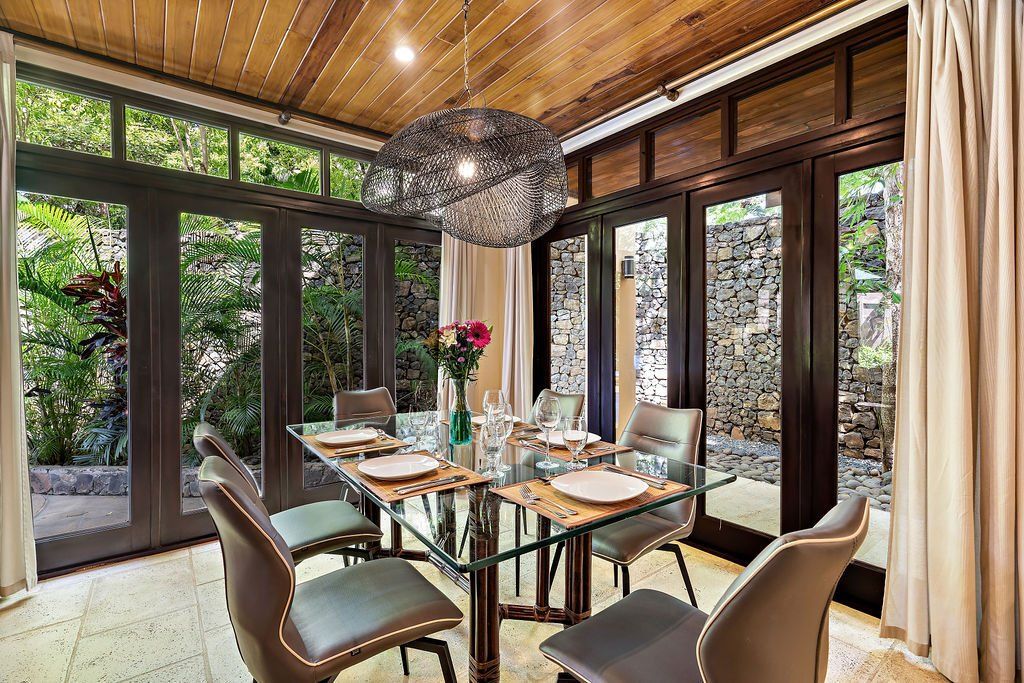 Modern dining room with glass table, beige chairs, wood ceiling, and large windows overlooking greenery