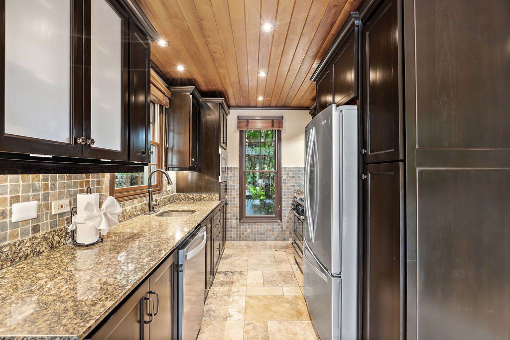 Narrow modern kitchen with dark cabinets, granite counters, stainless appliances, and wood plank ceiling.