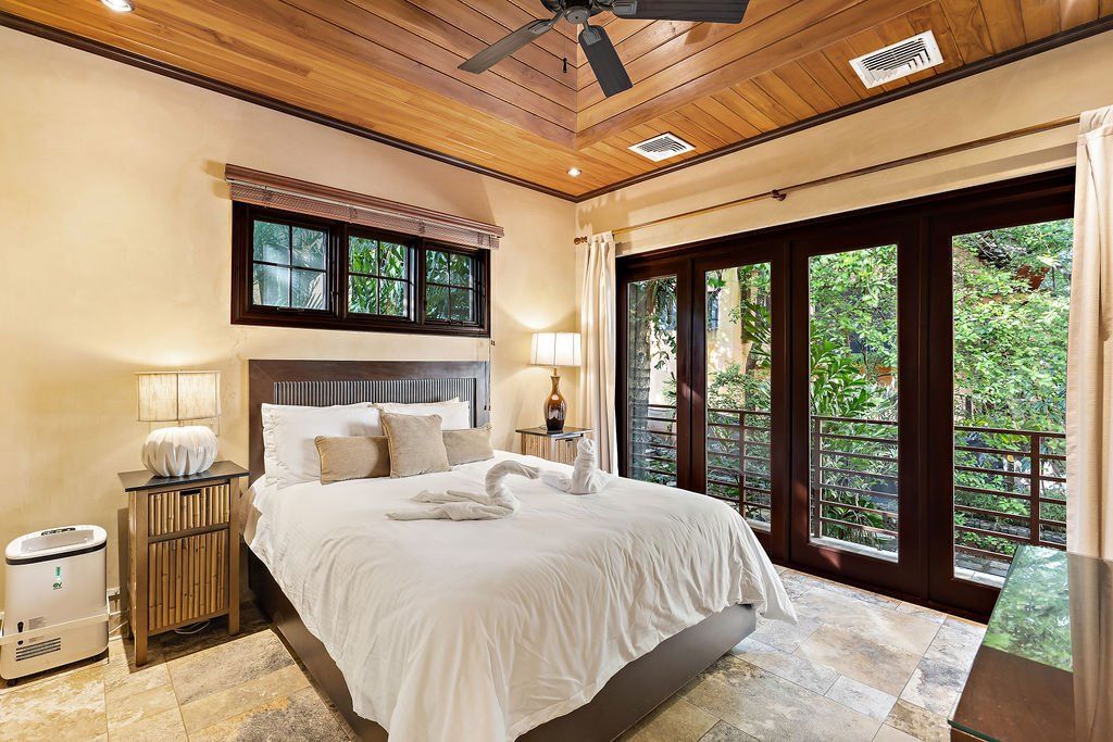 Bright bedroom with a large white bed, wood ceiling, and glass doors opening to greenery.