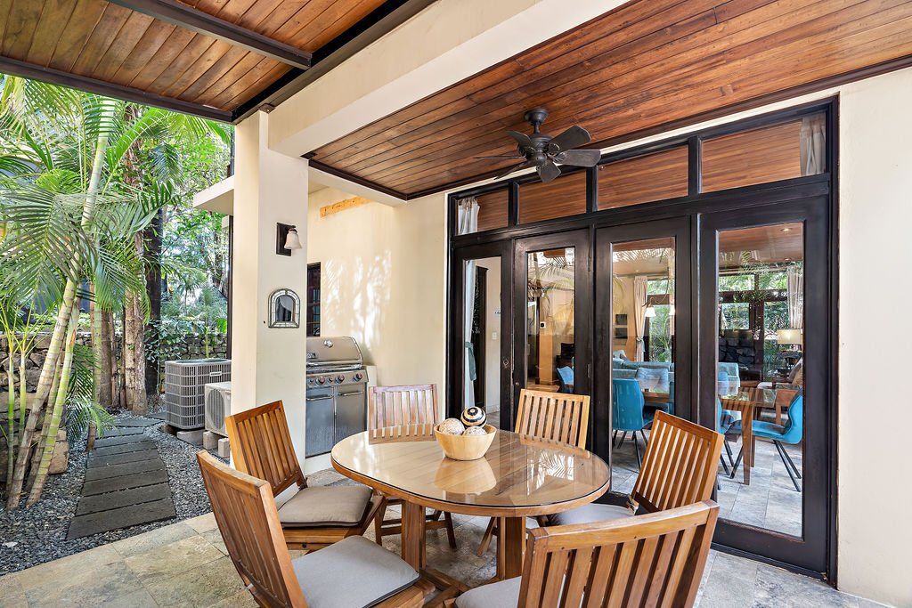 Covered patio with wooden table and chairs beside glass doors and garden greenery