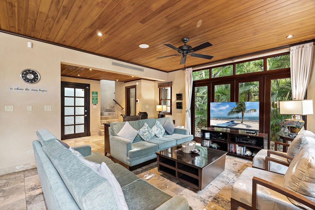 Bright living room with vaulted wood ceiling, light sofas, glass coffee table, and large windows overlooking greenery