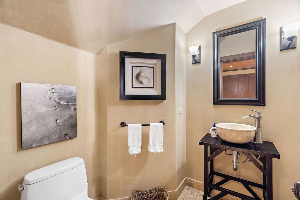 Small beige bathroom with toilet, wall art, towel rack, sink, mirror, and wall sconces.