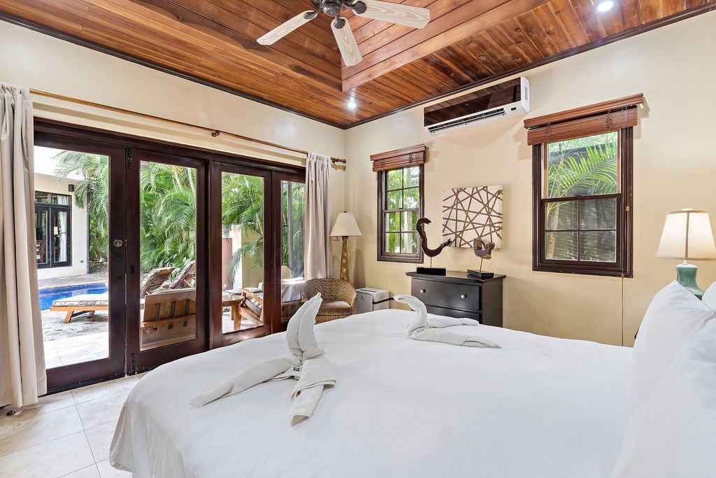 Bright tropical bedroom with white bed, wood ceiling, French doors, and a view of the pool outside