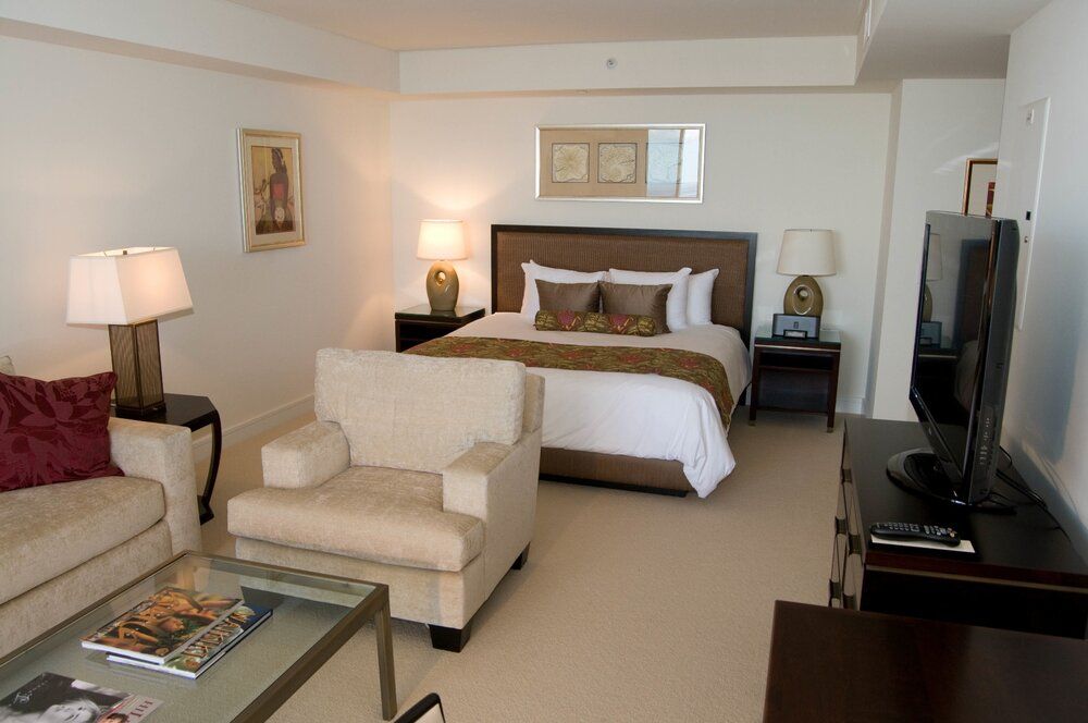 Modern hotel room with bed, armchair, TV, lamps, and neutral decor