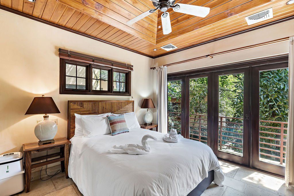 Bright bedroom with a white bed, wooden ceiling, and large windows opening to greenery.