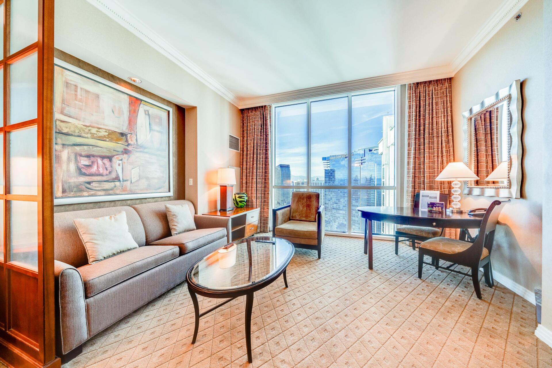 Bright living room with sofa, glass coffee table, desk, and floor-to-ceiling windows overlooking city buildings
