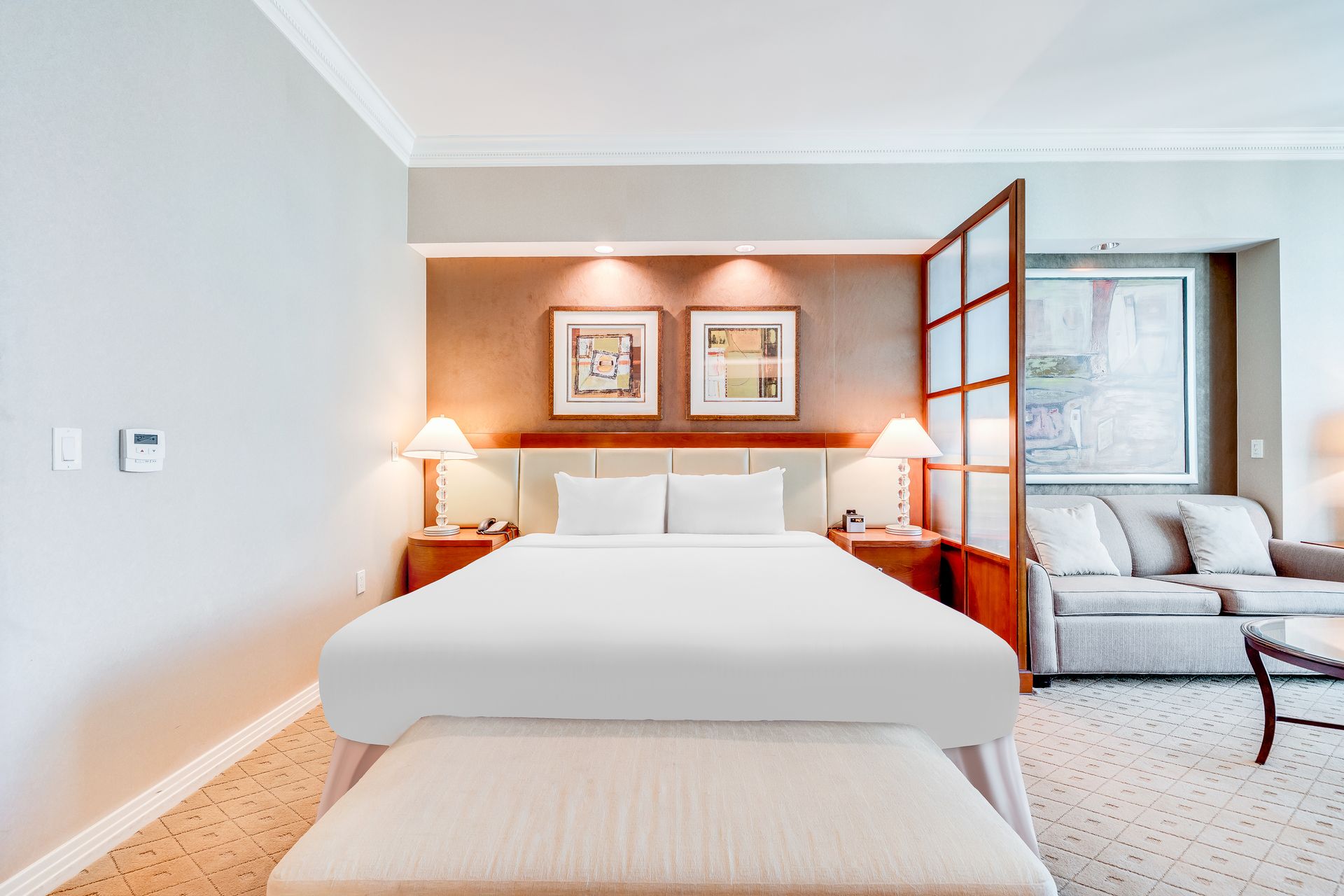 Bright hotel bedroom with a white bed, warm bedside lights, framed art, and an adjoining sitting area
