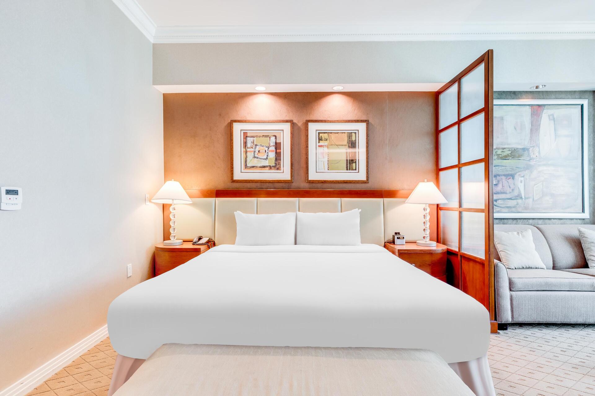 Modern bedroom with white bed, wood headboard, bedside lamps, and glass partition to a bright sitting area