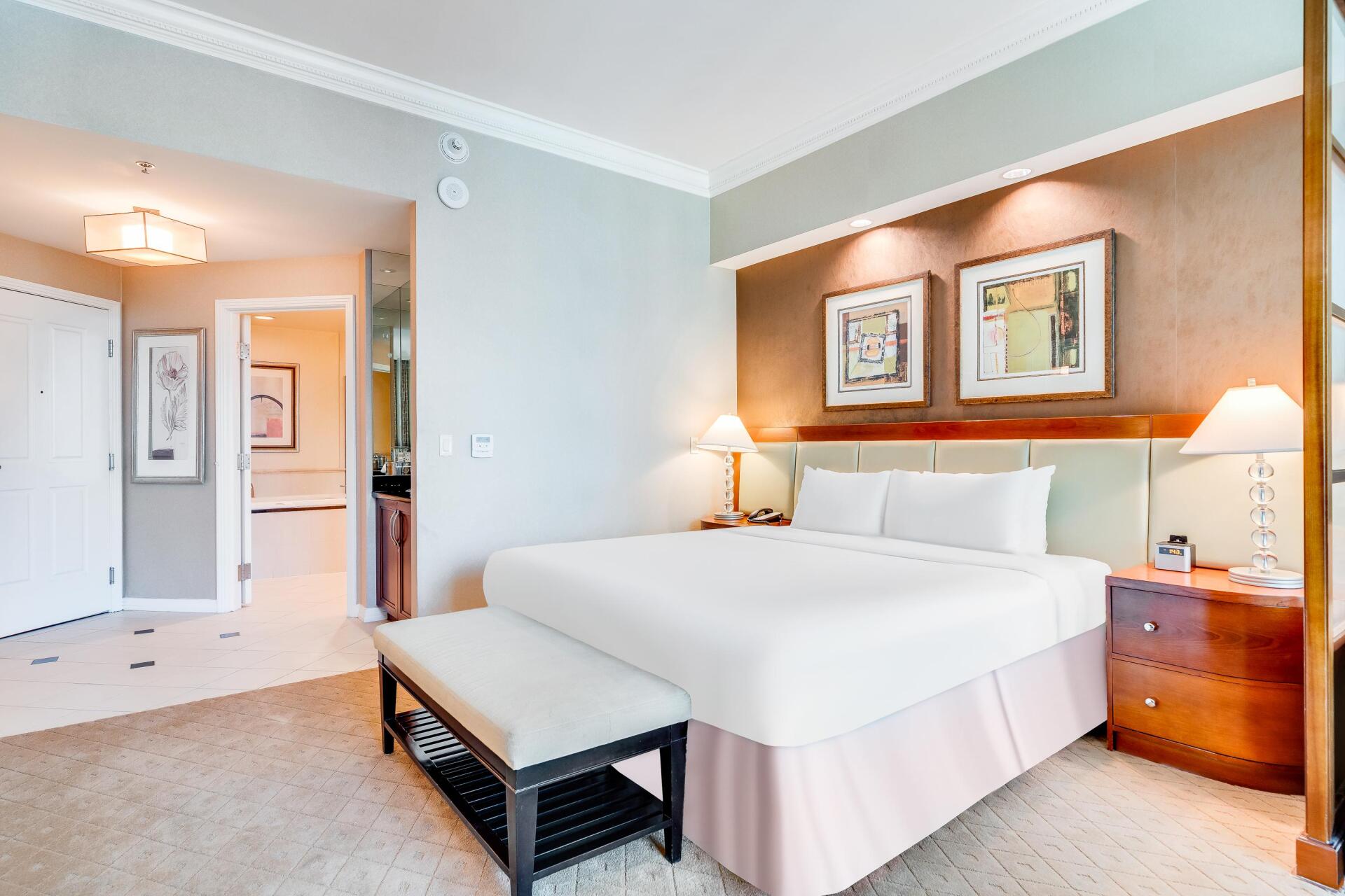 Bright bedroom with a made bed, bedside lamps, and a bench, viewed from the doorway