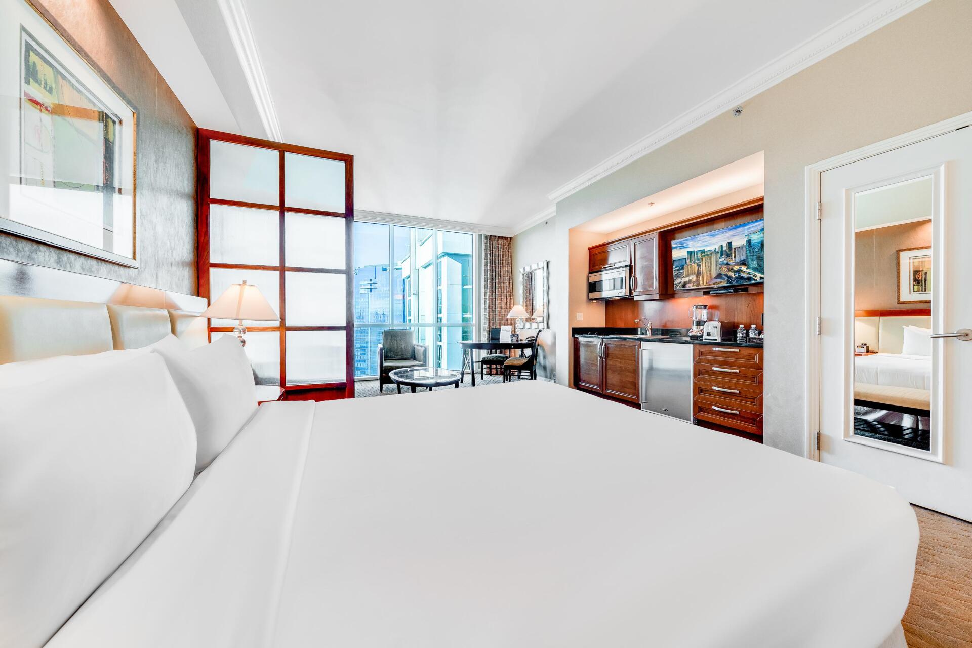 Bright modern bedroom with white bedding, open glass doors, and a view into a kitchen area