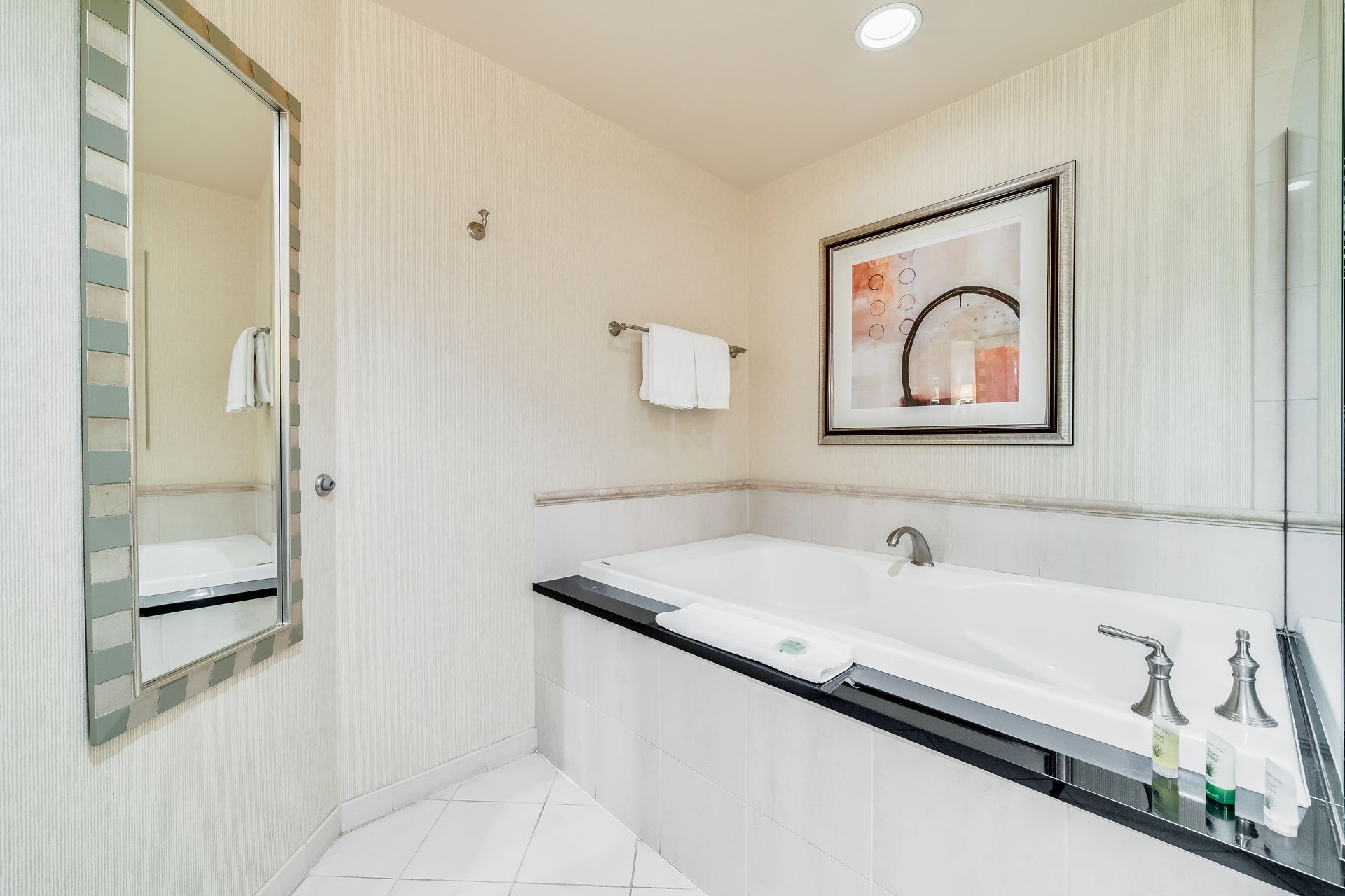 Bright bathroom with a large tub, wall mirror, framed art, and white tiled walls
