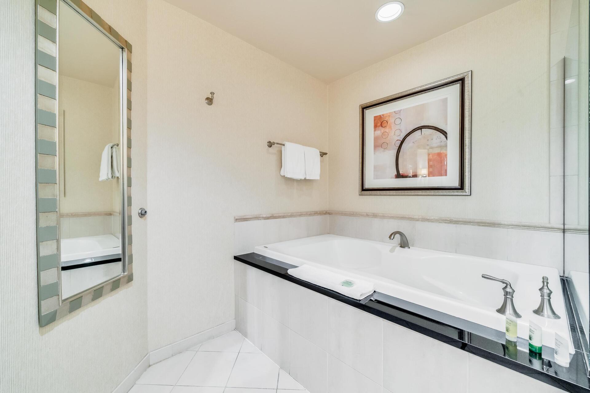 Bright modern bathroom with white tile, large soaking tub, wall mirror, and framed artwork