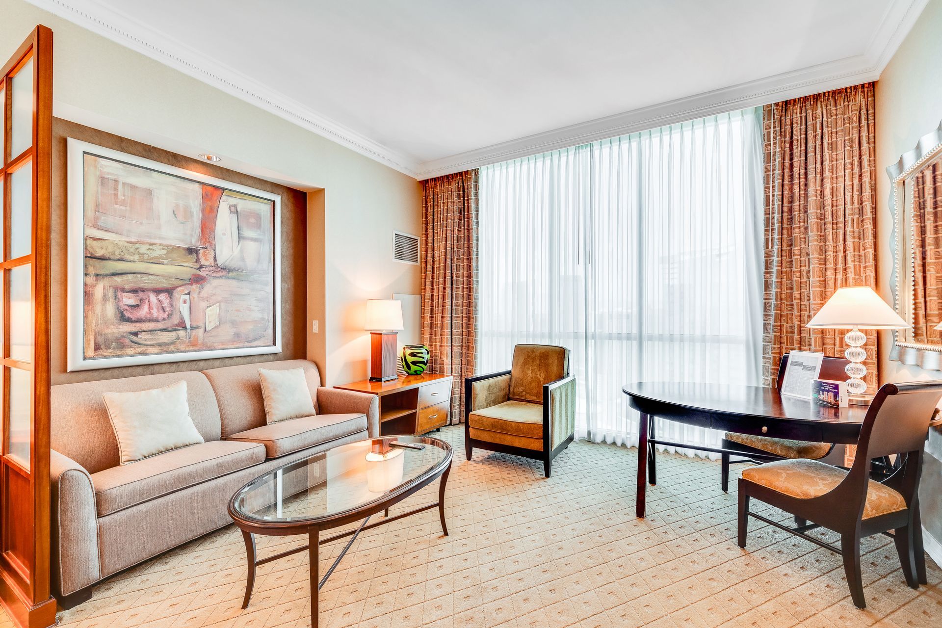 Bright hotel-style living room with beige sofa, glass table, armchairs, and large window with curtains