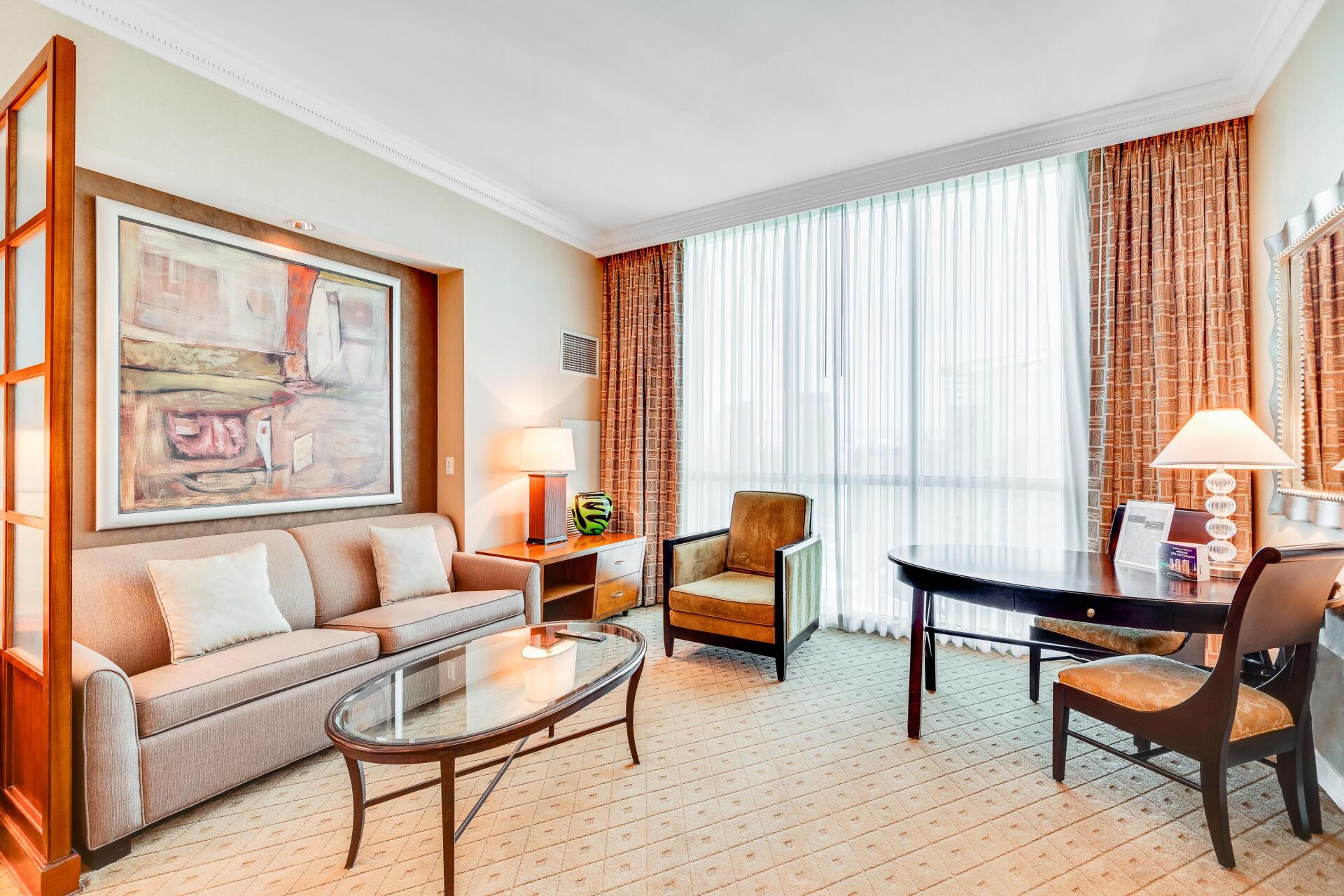 Bright living room with beige sofa, glass coffee table, armchair, desk, and large sheer-curtained window
