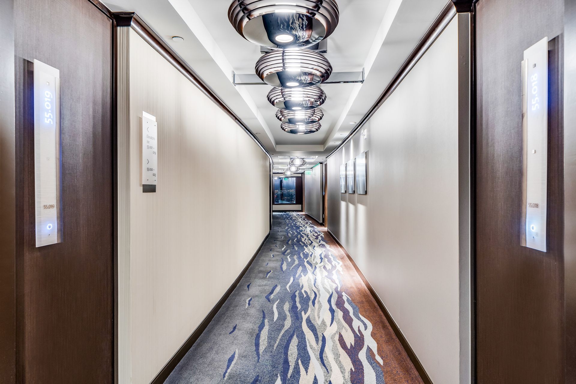 Modern hotel hallway with wood-paneled walls, blue patterned carpet, and ceiling lights