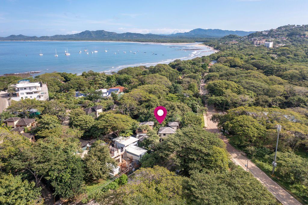 Aerial view of a tropical coastline with dense green trees, blue water, and scattered buildings.