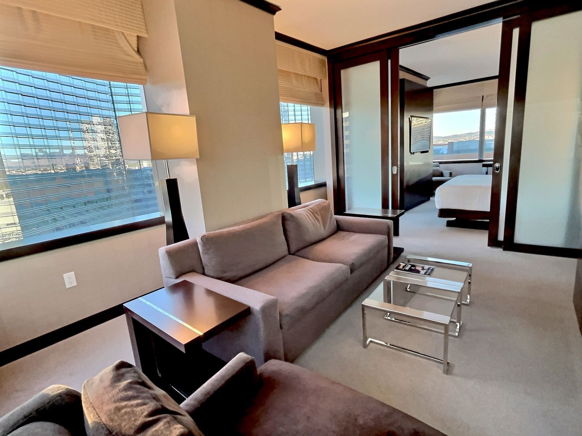 Modern living room with gray sofa, glass tables, floor lamp, and bedroom visible through open doorway.