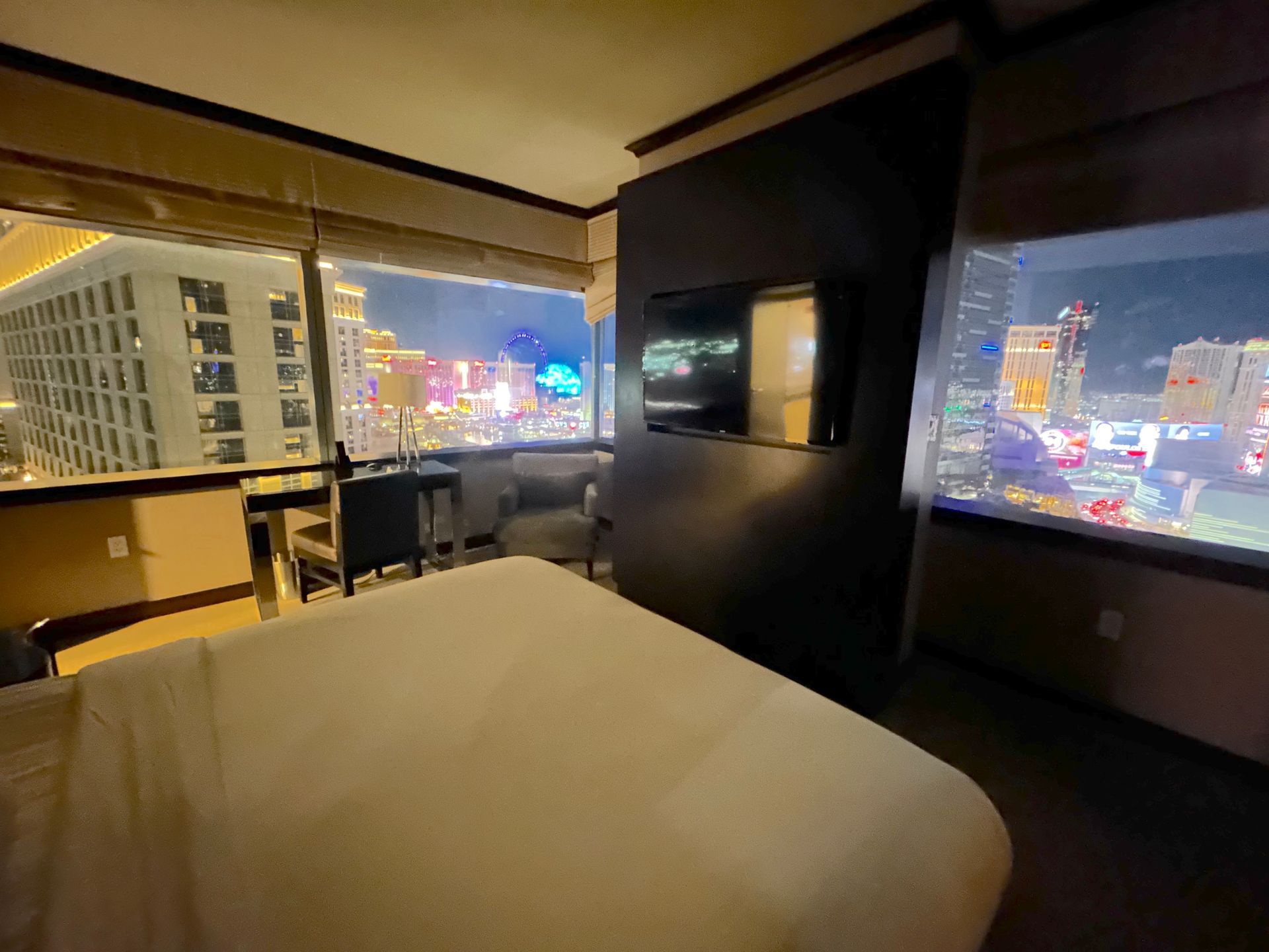 Hotel room with city skyline views at night, a bed in the foreground and large windows on two sides.