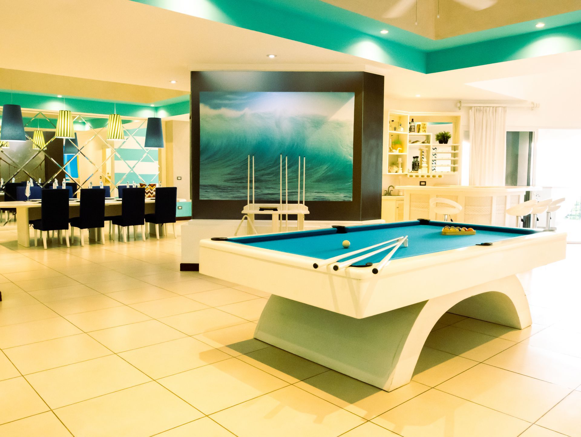 Modern game room with a pool table, bar stools, and ocean-themed decor, lit by teal and white lights