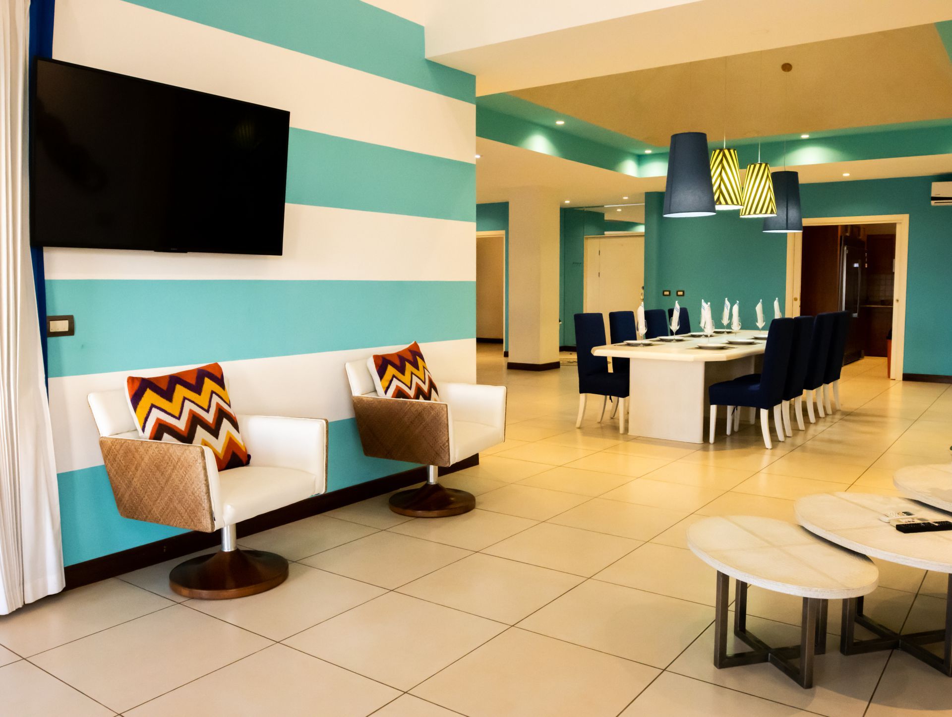 Modern living room with teal-striped walls, white chairs, dining table, and wall-mounted TV