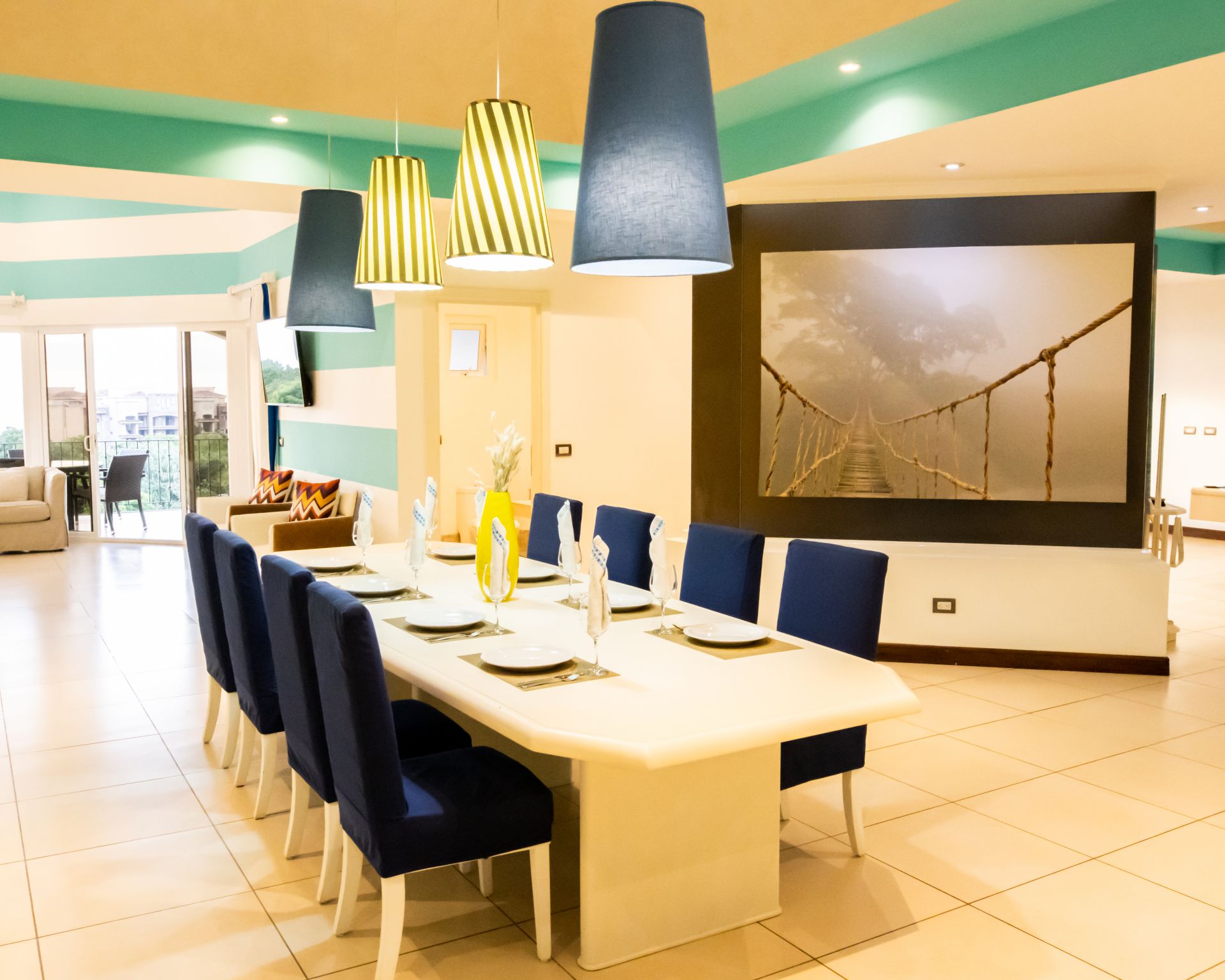 Modern dining room with a long white table, navy chairs, and colorful pendant lights in a bright interior.