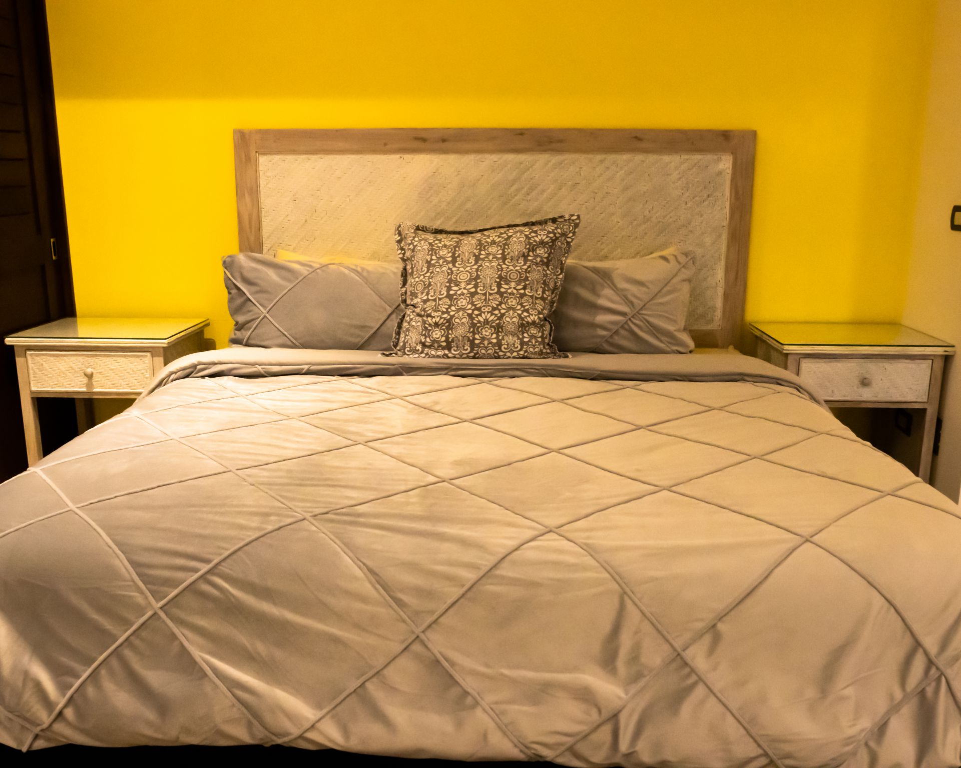 Neatly made bed with beige quilt and patterned pillows against a bright yellow wall, flanked by two nightstands.
