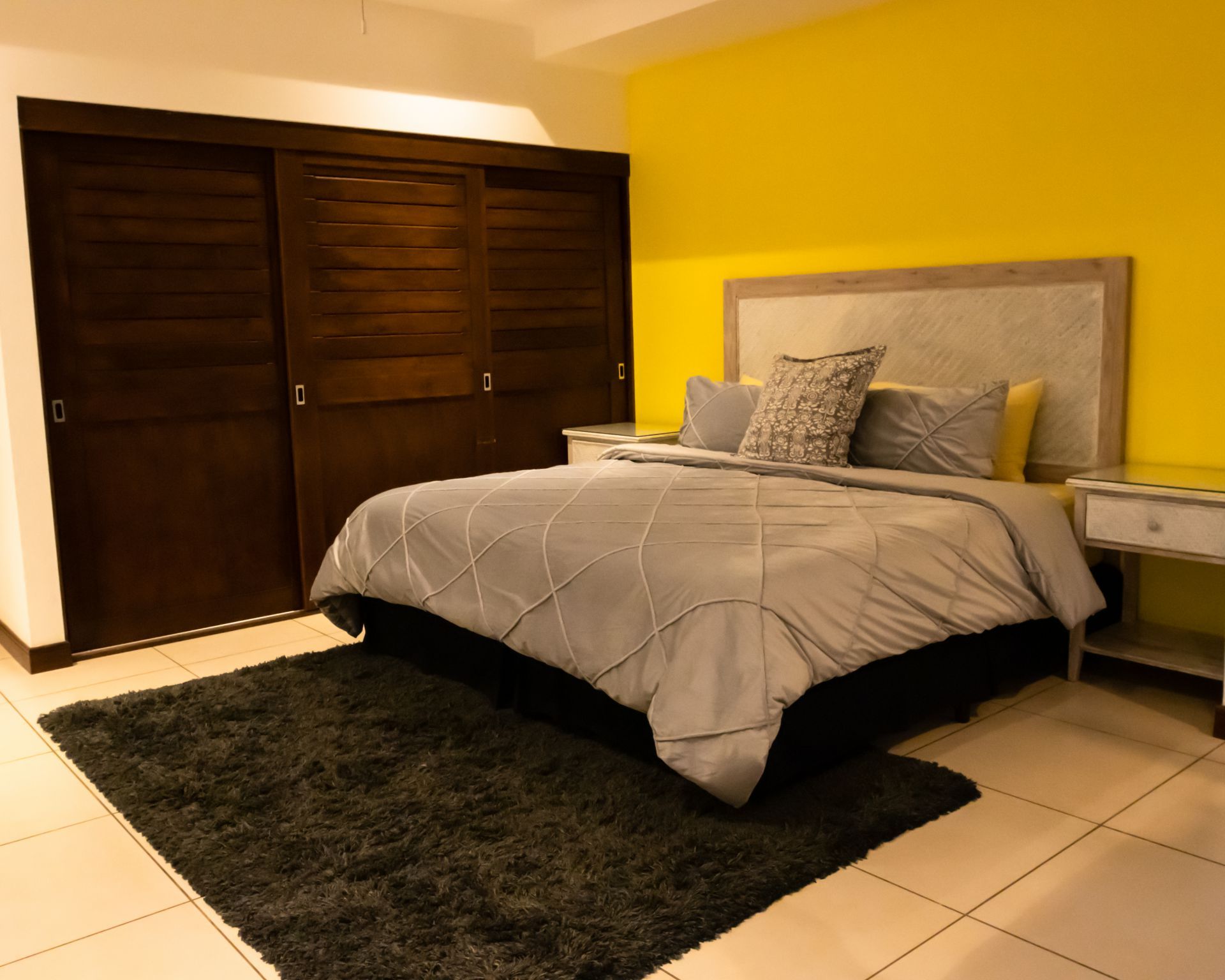 Modern bedroom with a gray bed, dark wardrobe, black rug, and yellow accent wall