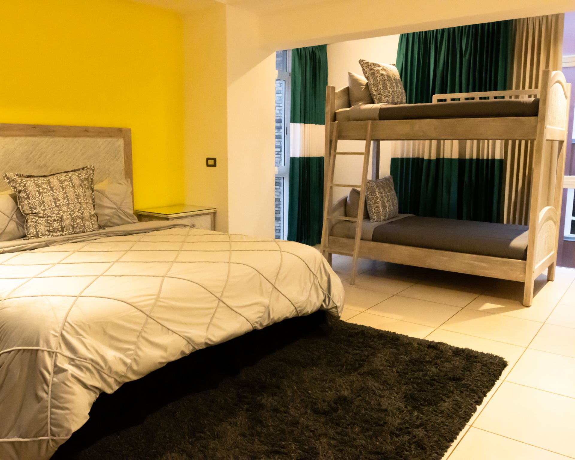 Bedroom with beige bed, bunk bed, yellow wall, green curtains, and dark rug.