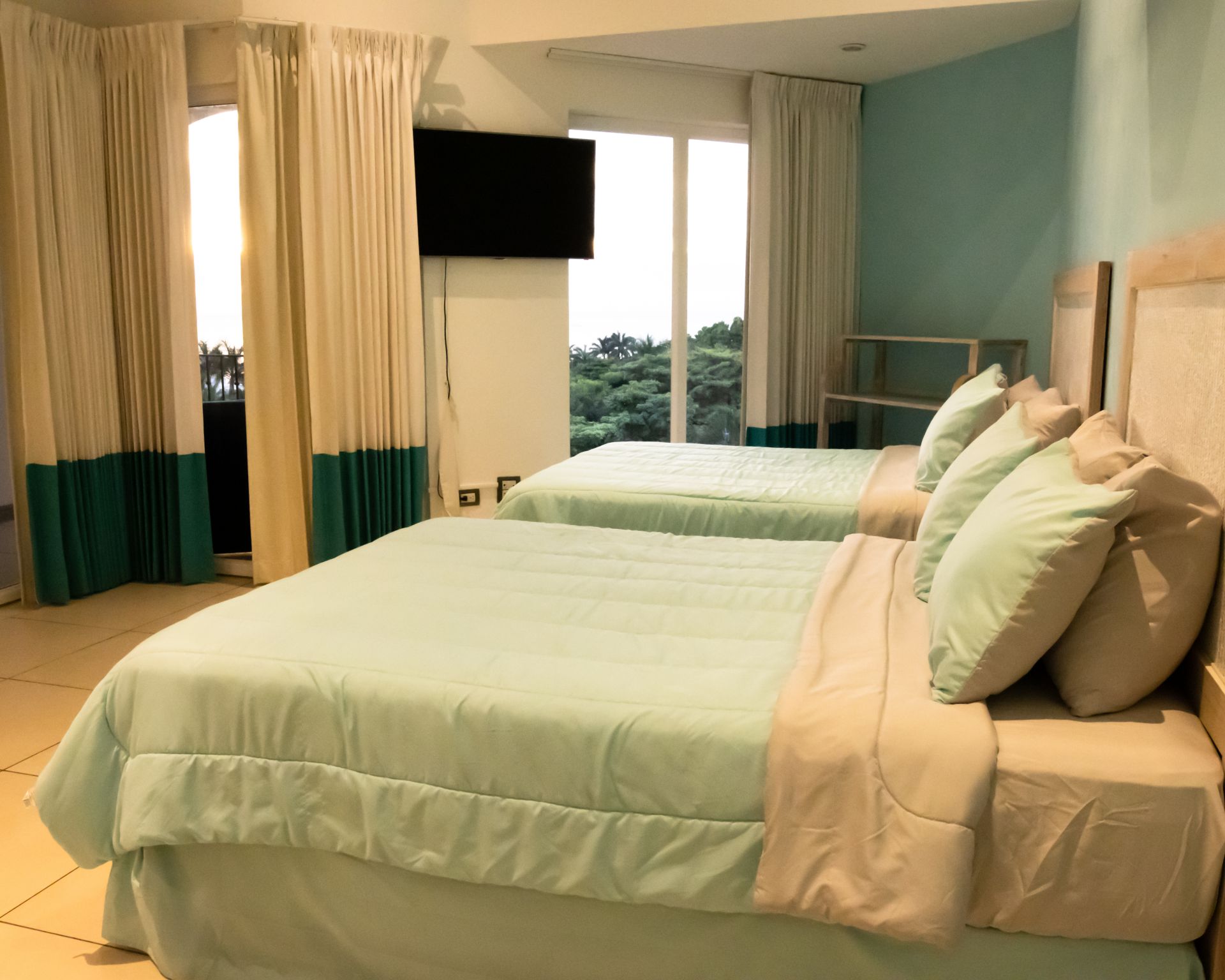 Hotel room with two neatly made beds, curtains, wall-mounted TV, and ocean view through the window