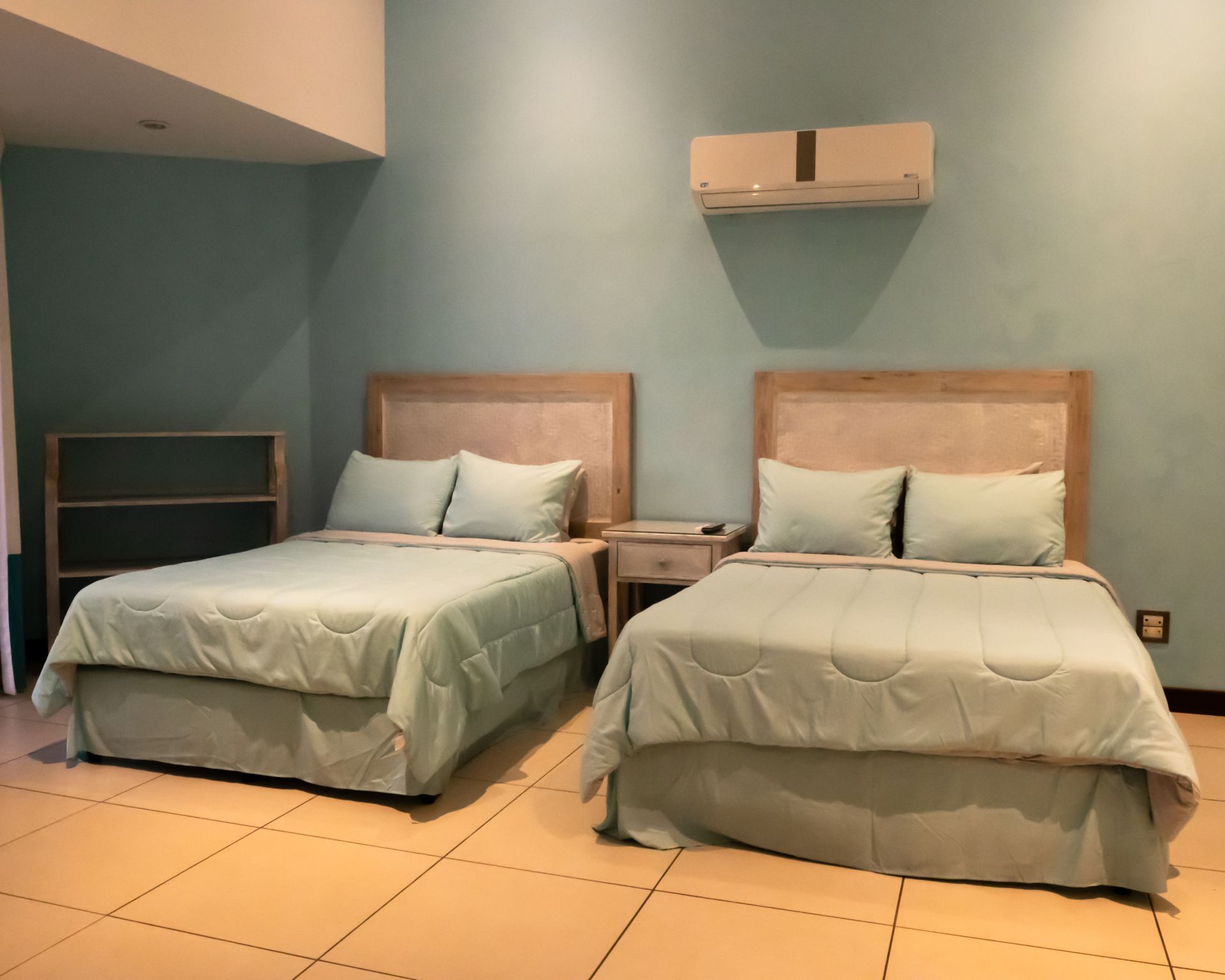 Two twin beds in a teal hotel room with beige tile floor and a wall-mounted air conditioner