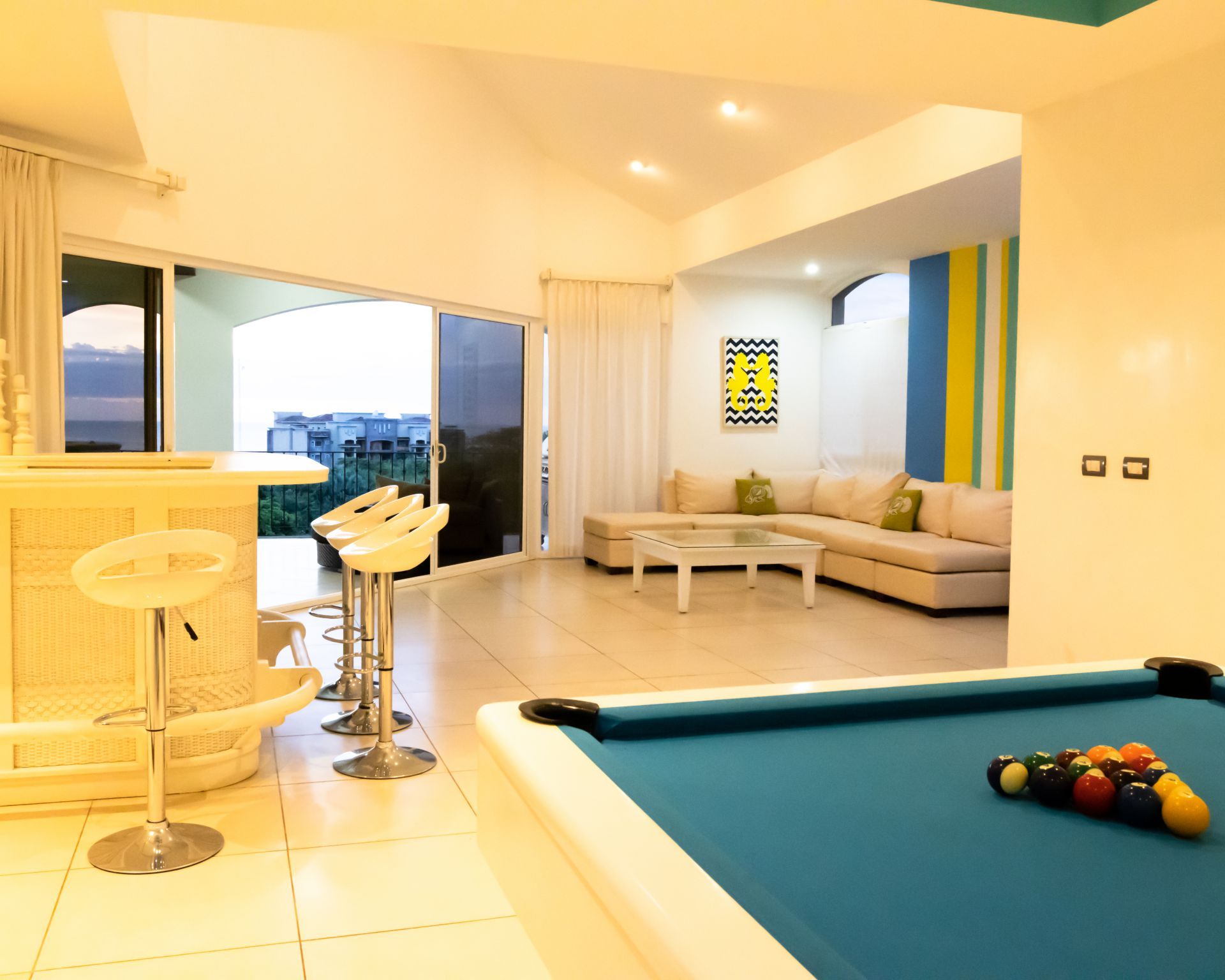Modern living room with pool table, bar stools, sofa, and large windows overlooking a sunset view.