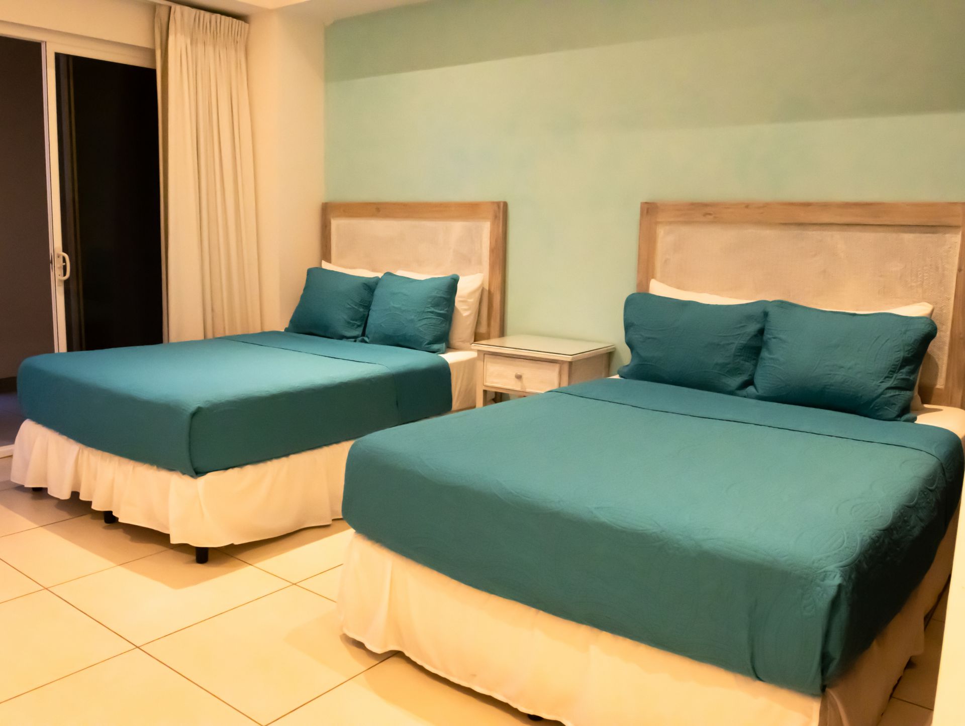 Two teal-covered beds with white skirts in a softly lit bedroom with cream walls and tiled floor