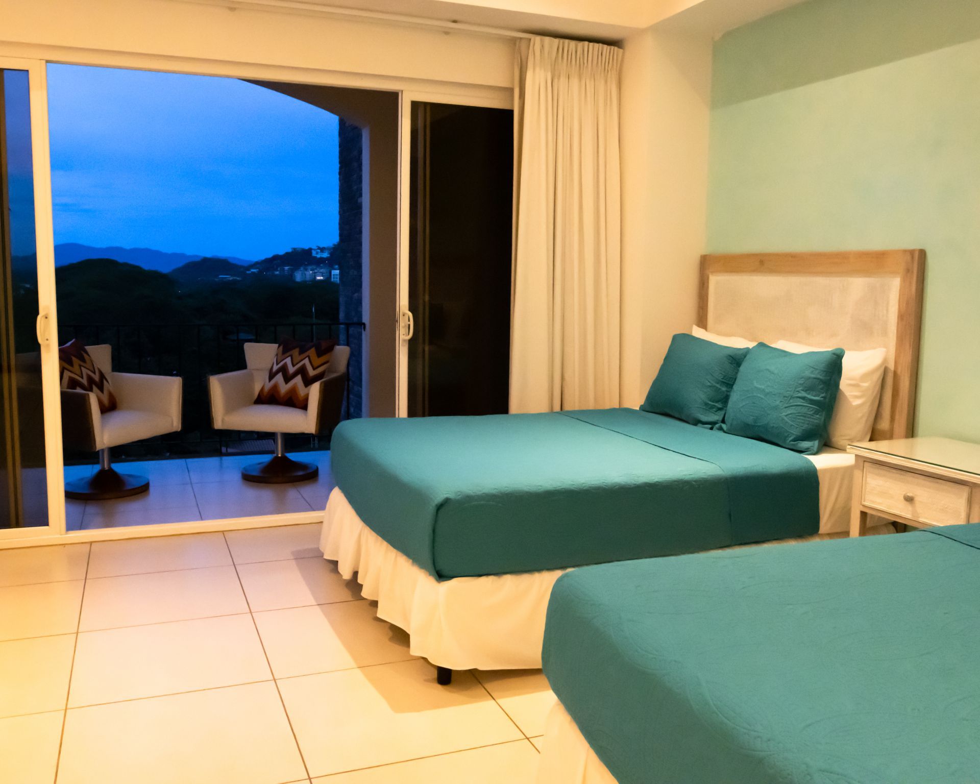 Bright bedroom with two teal beds, white tile floor, and sliding doors to a sunset balcony.