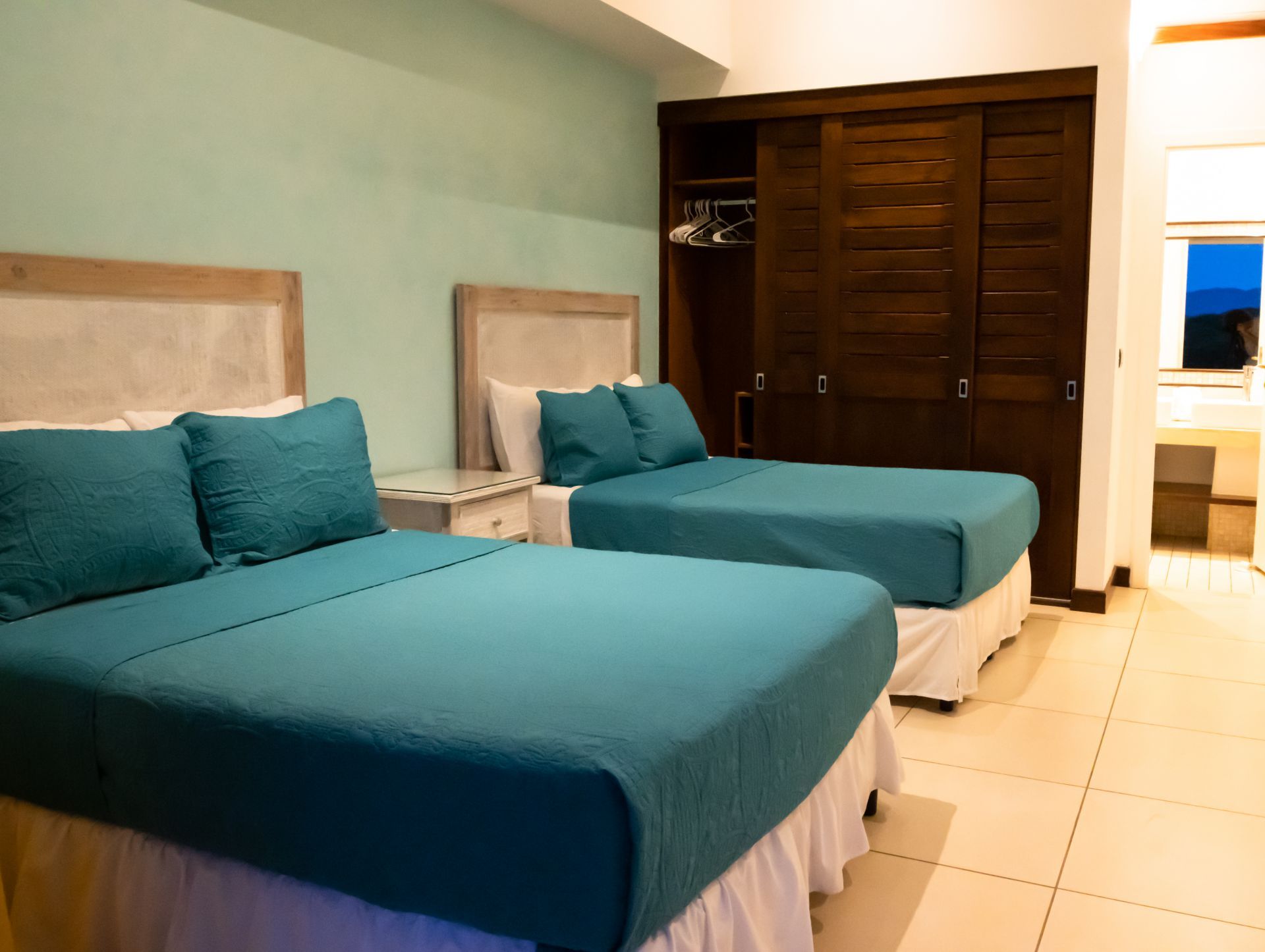 Two teal beds in a bright hotel room with a large wooden wardrobe and tiled floor