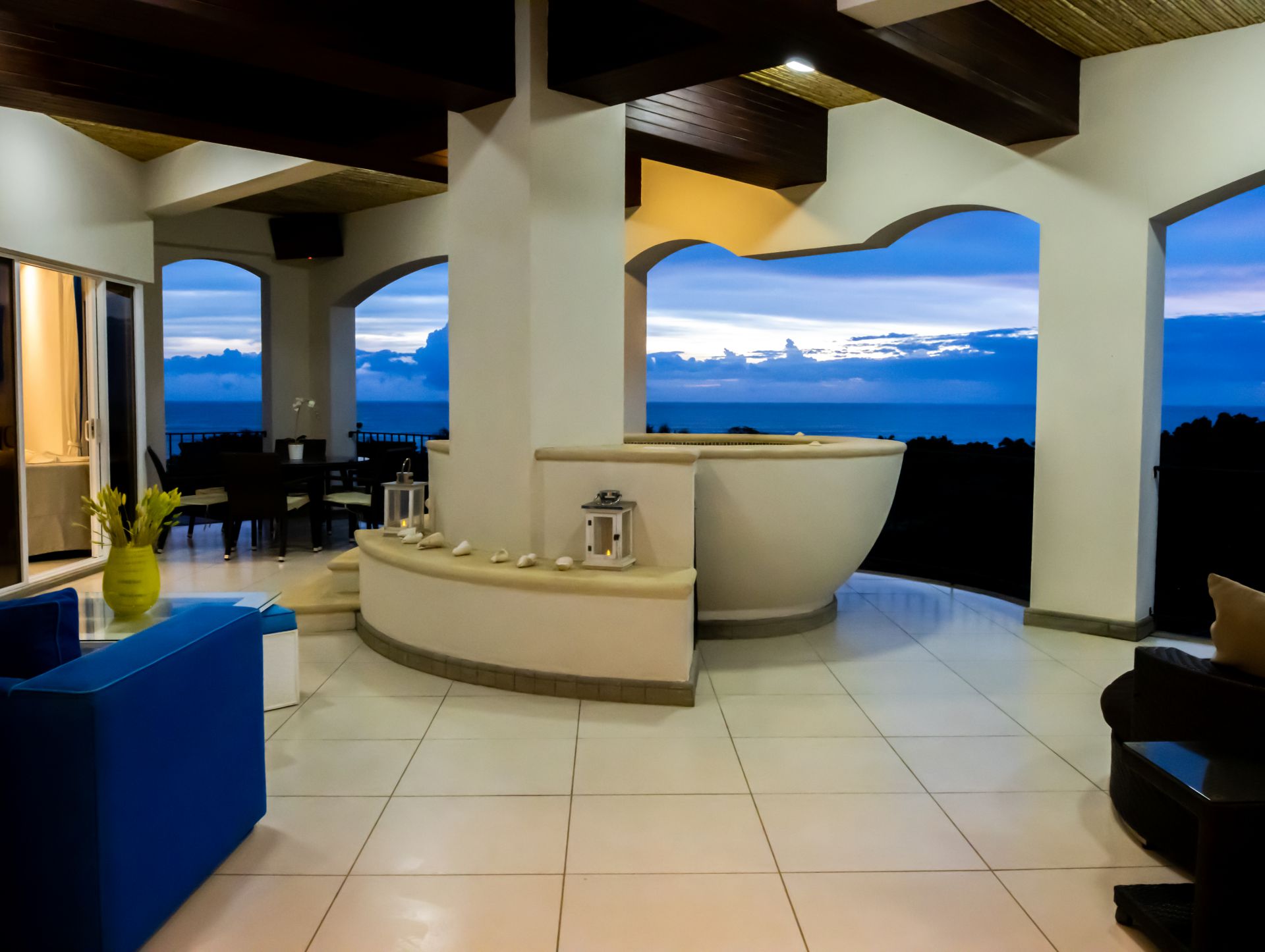 Open-air luxury bathroom with tiled floor, curved tub, and ocean sunset view