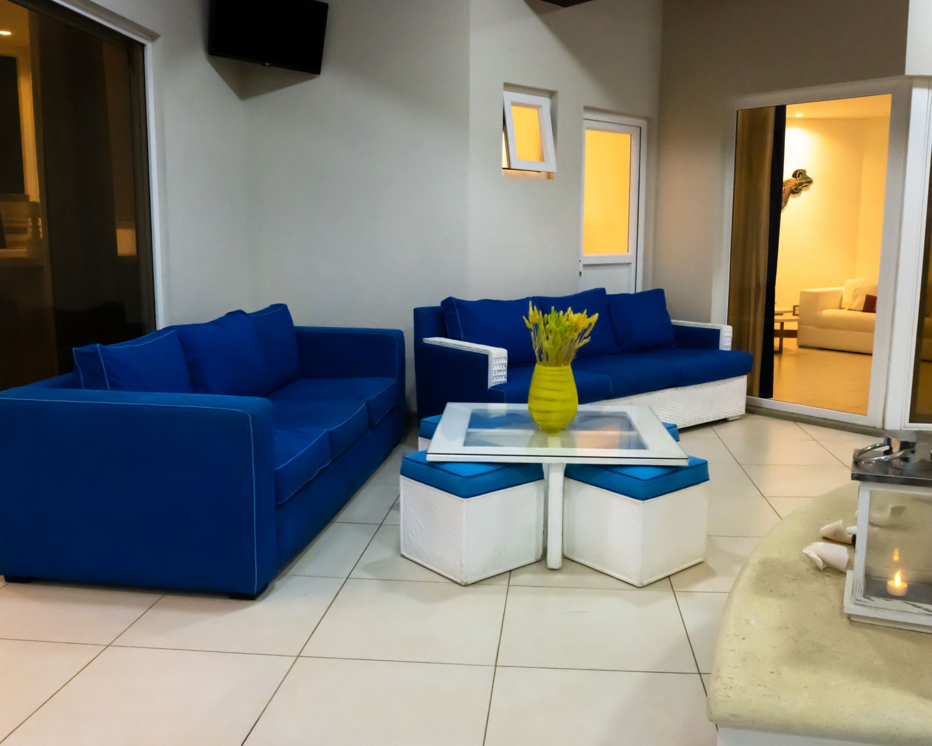 Modern living room with blue sofas, white glass coffee table, and open doorway to a lit bedroom.
