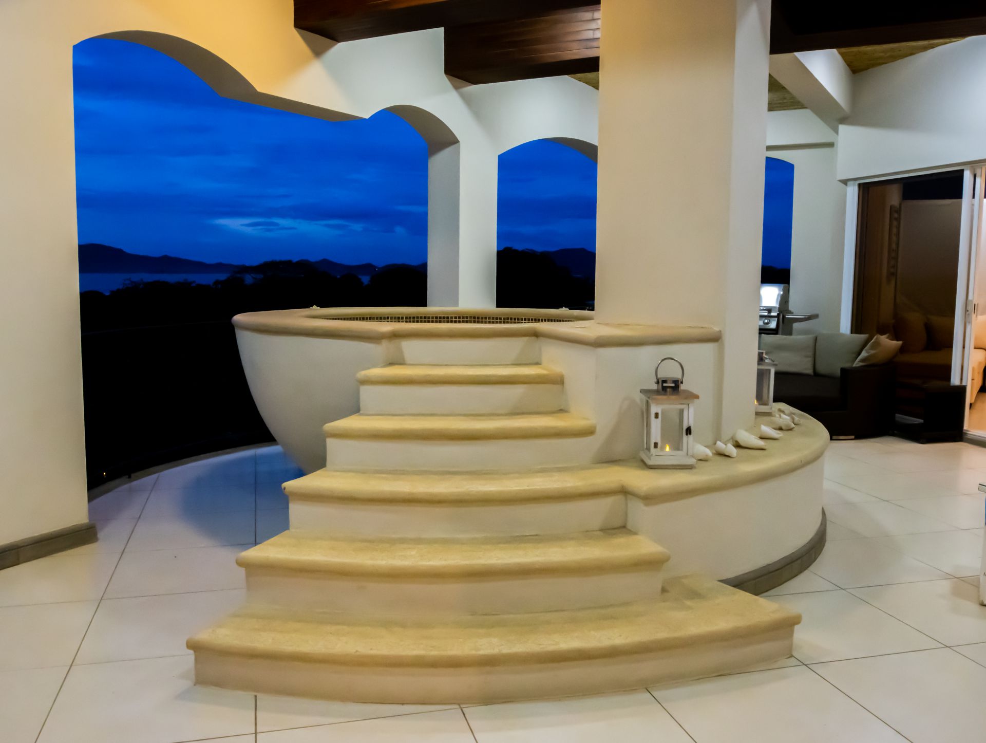 Spiral stone stairs leading to an indoor/outdoor pool at dusk, with columns and a lantern on the right