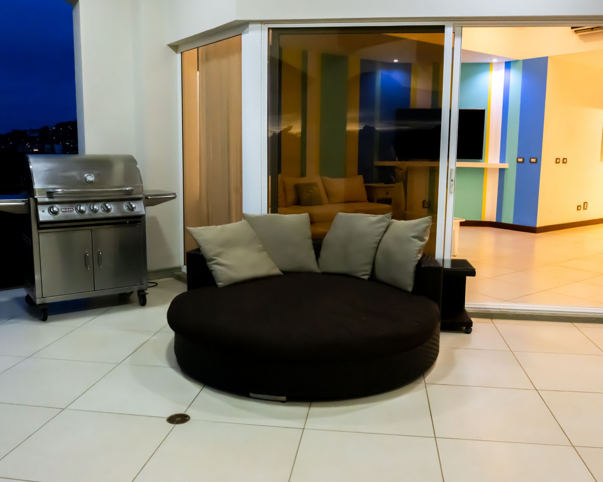 Modern patio with round black sofa and grill beside glass sliding doors at dusk