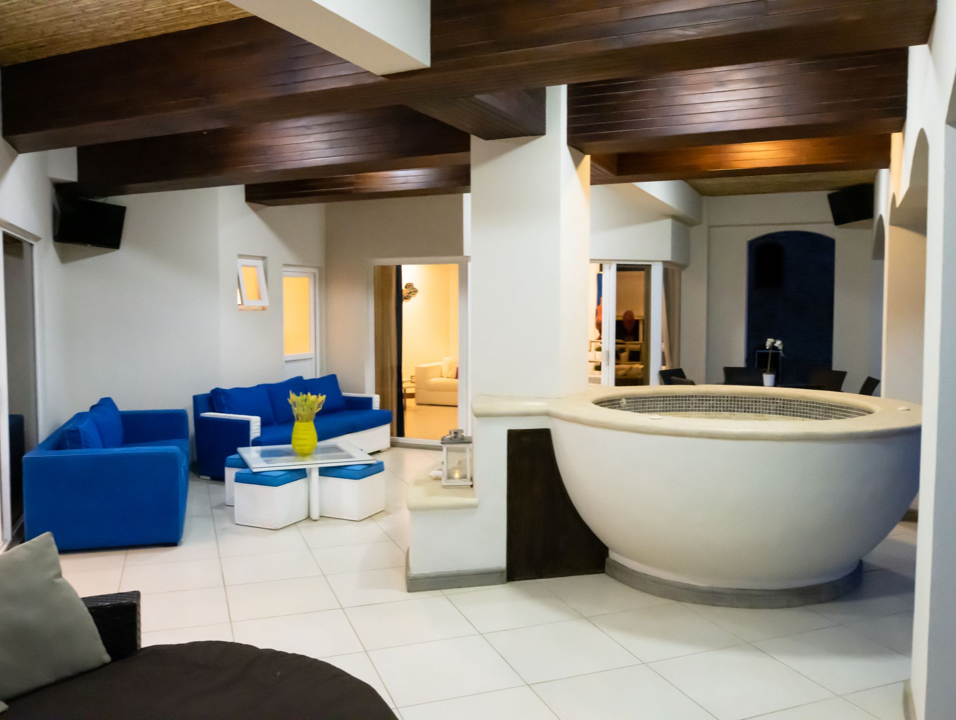 Modern living room with blue sofas, white tile floor, and a large round bathtub by a wooden beam ceiling