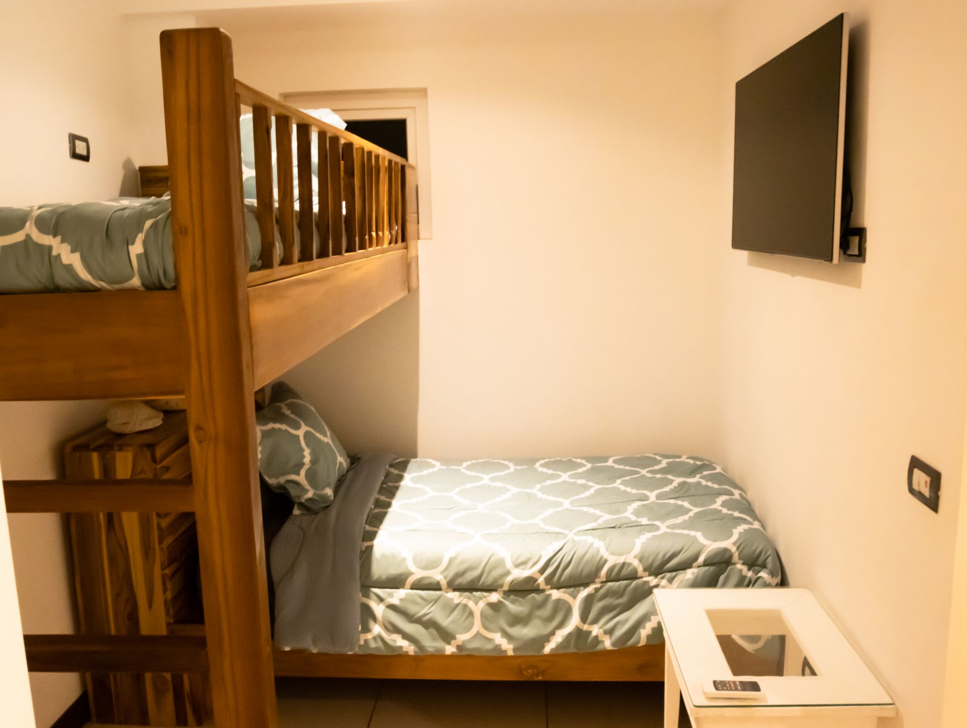 Small bedroom with wooden bunk bed, gray patterned bedding, wall TV, and bedside table.