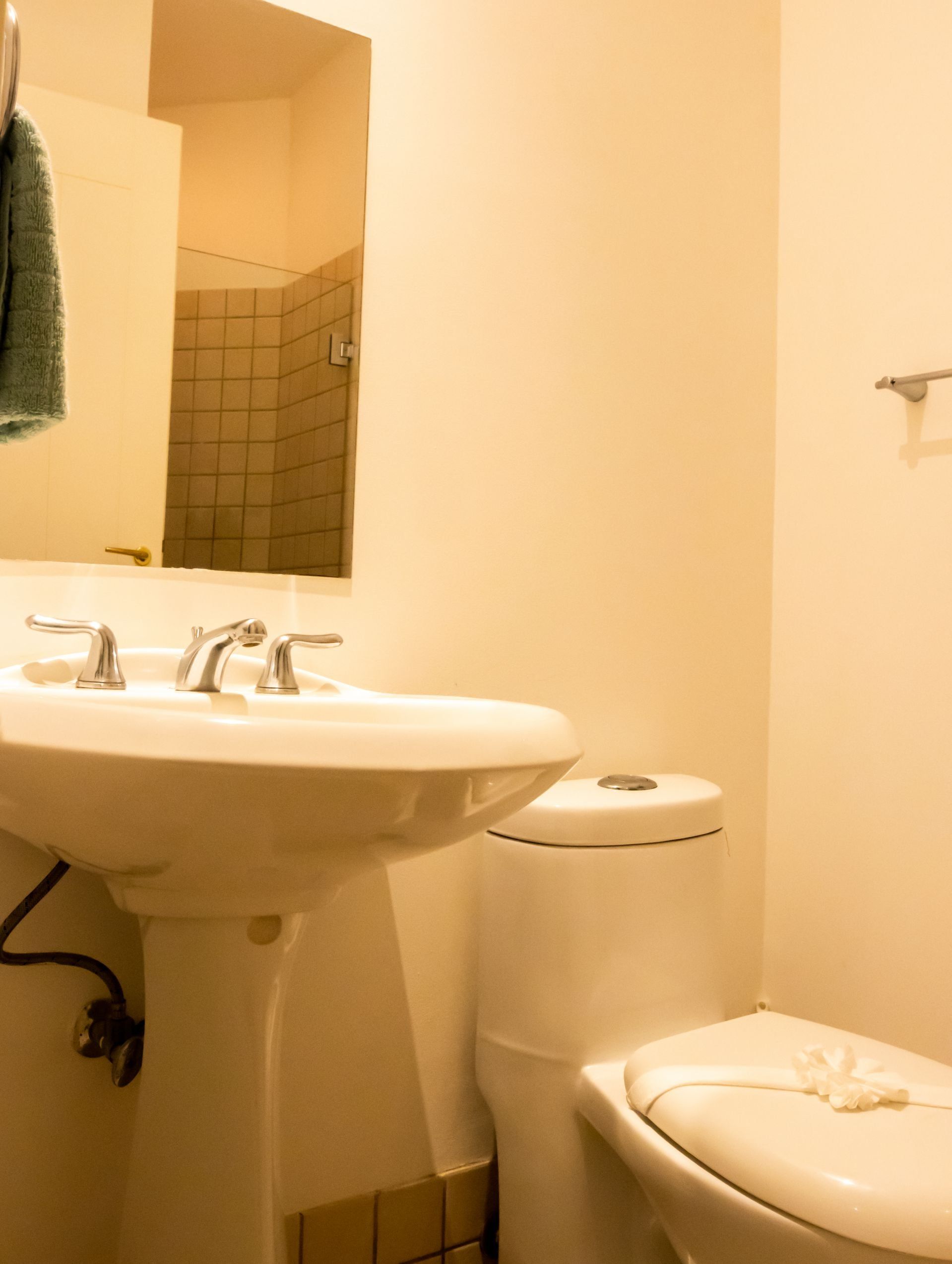 Small beige bathroom with pedestal sink, toilet, mirror, and shower visible in reflection