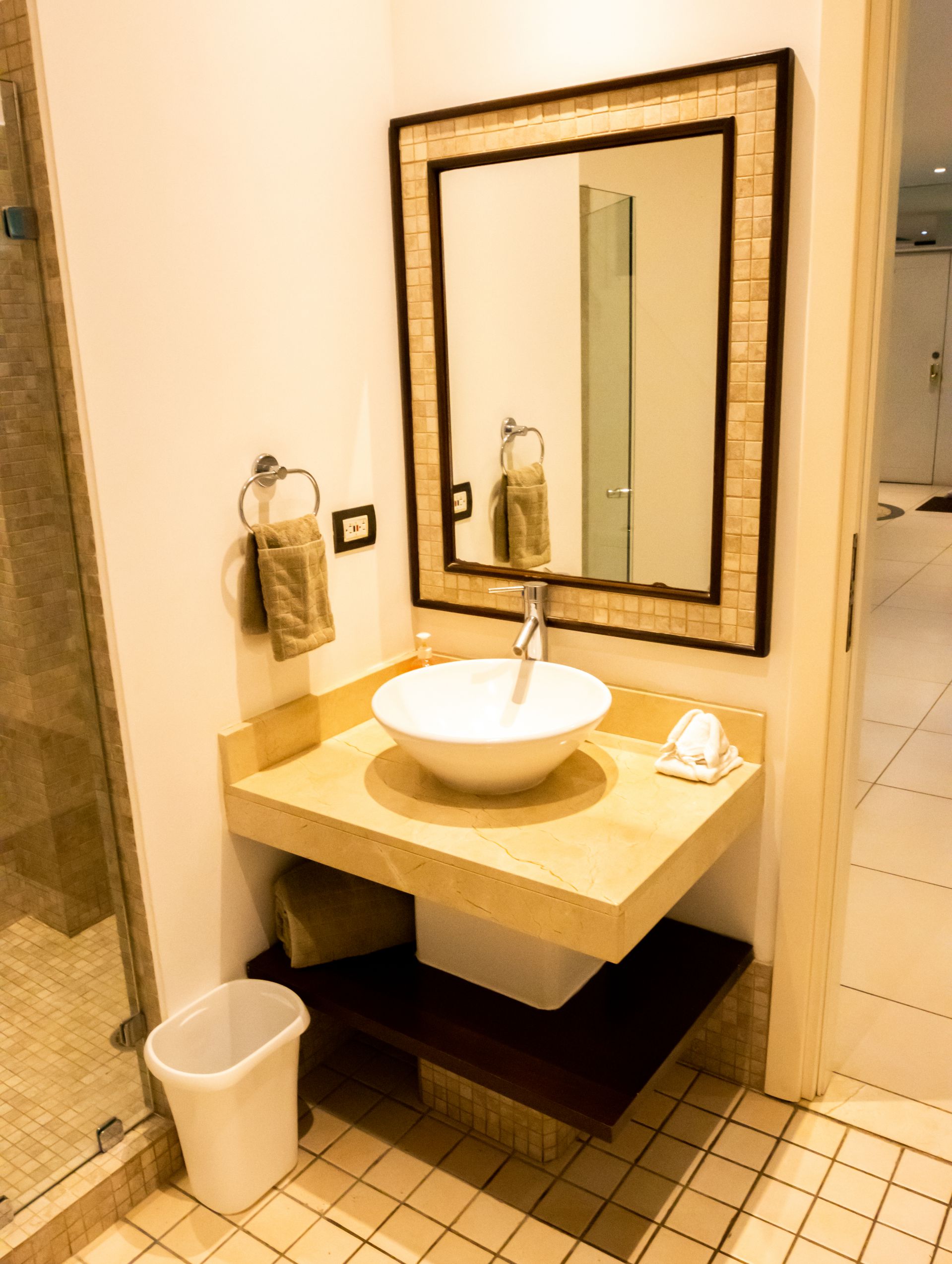 Small bathroom vanity with vessel sink, mirror, towel ring, and trash bin on tiled floor.