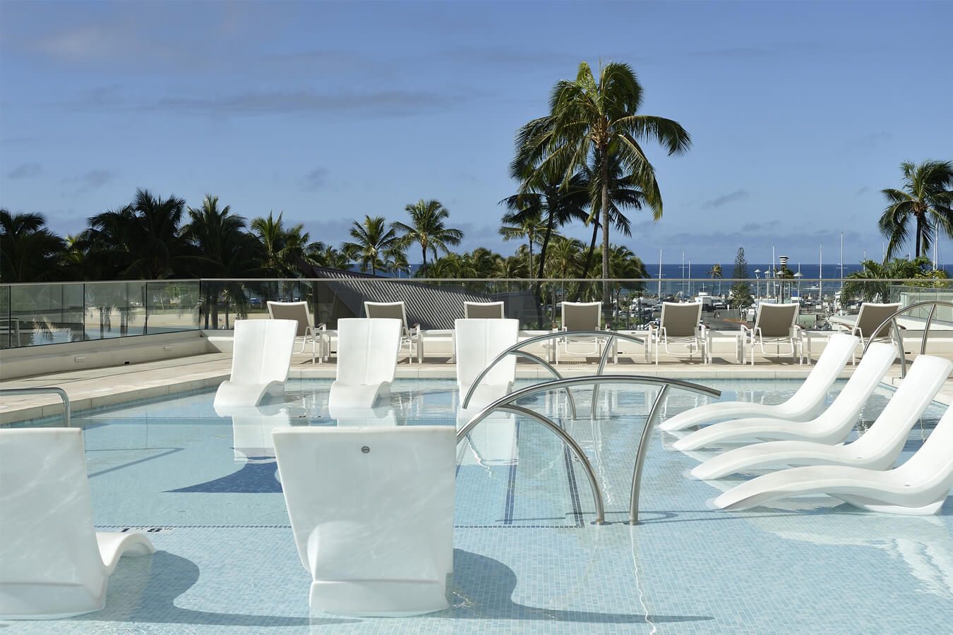 Poolside lounge chairs beside a shallow resort pool with ocean view and palm trees