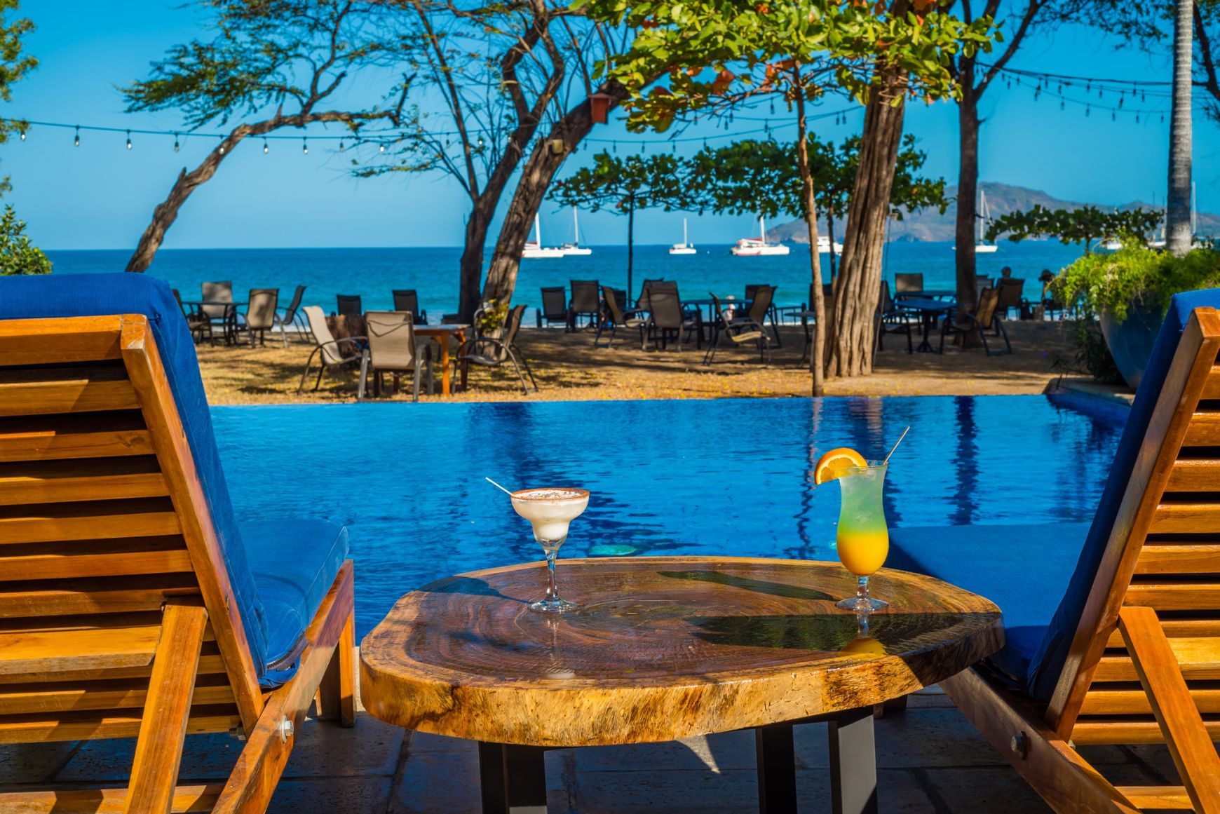 Resort poolside view with lounge chairs, ocean, and two cocktails on a table at sunset